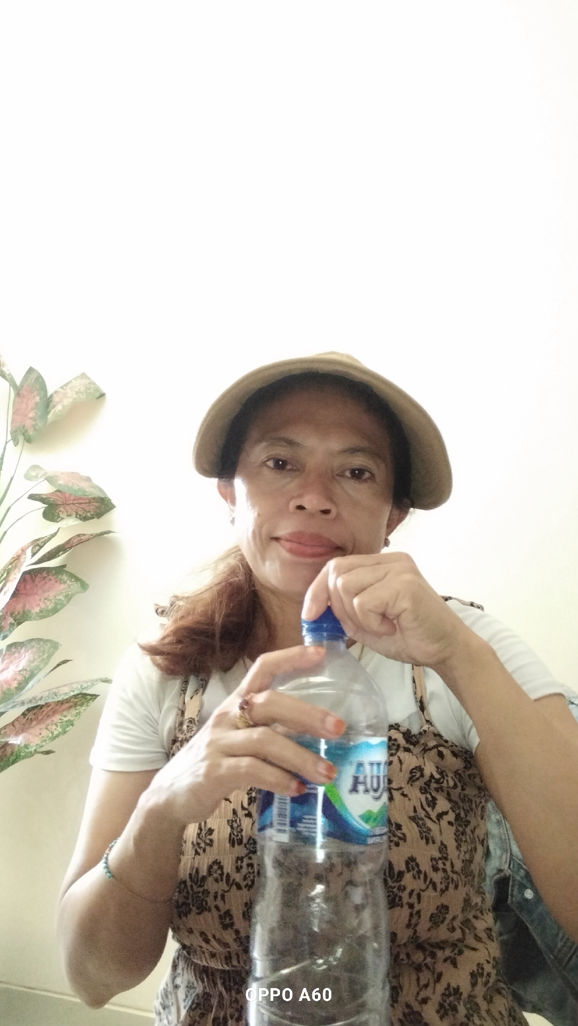 A medium shot shows a woman from the chest up, wearing a light-colored t-shirt, a patterned dress, and a light brown hat. She has medium-length, dark brown hair and is looking towards the camera with a slight smile, her tongue slightly out. Her right hand is holding the cap of a clear plastic water bottle, and her left hand is near the bottle. A plant with large, colorful leaves is visible to the left of the woman. The background is a plain, light-colored wall. The bottom right corner of the image displays the text "OPPO A60". The overall scene appears to be indoors, likely in Denpasar, Indonesia, based on the visible text which may indicate the phone model used for the photograph.