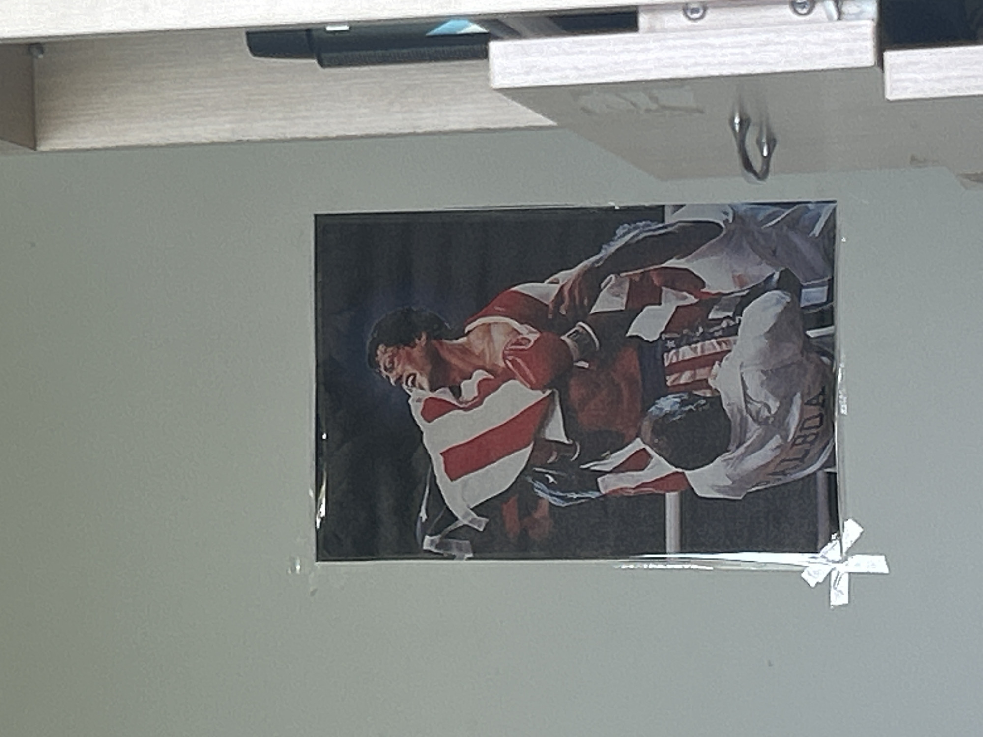 A rectangular photograph of two people in a possible athletic competition is taped to a gray wall with clear tape. One person is wearing a red and white striped jersey and black shorts, while the other is wearing a white jersey and black shorts with white stars. The background of the photograph is a dark gray or black color. The location is unknown, Unknown.