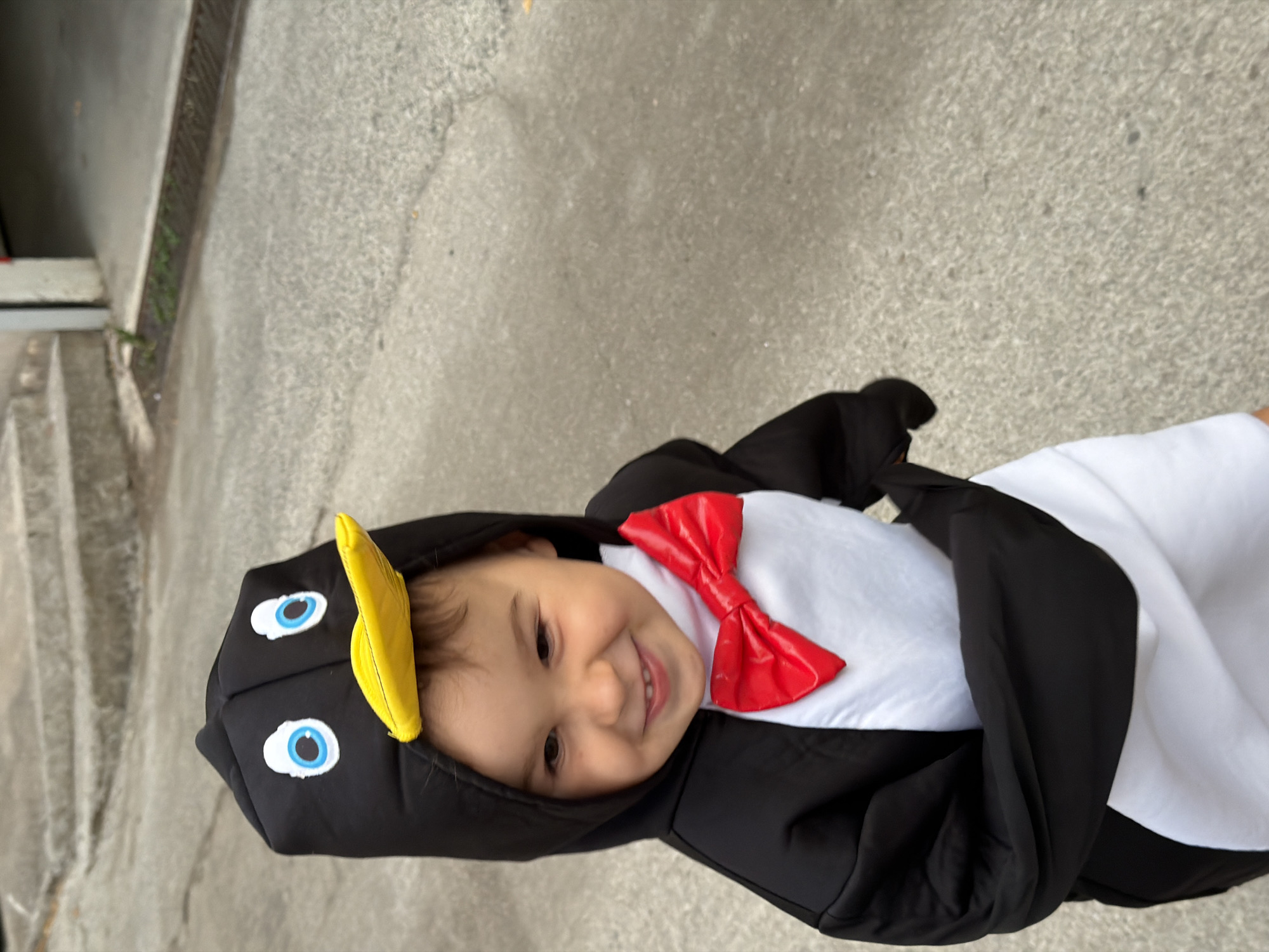 A smiling young child, dressed in a penguin costume featuring a black body, white belly, red bow tie, yellow beak, and blue eyes, is captured outdoors on a concrete surface. The setting appears to be in an urban environment, possibly in the city of Budva, Montenegro, considering the visible concrete and building structures in the background. The child's smile is prominent, and the costume is detailed with recognizable penguin features. The composition is a close-up shot, focusing on the child and the costume.