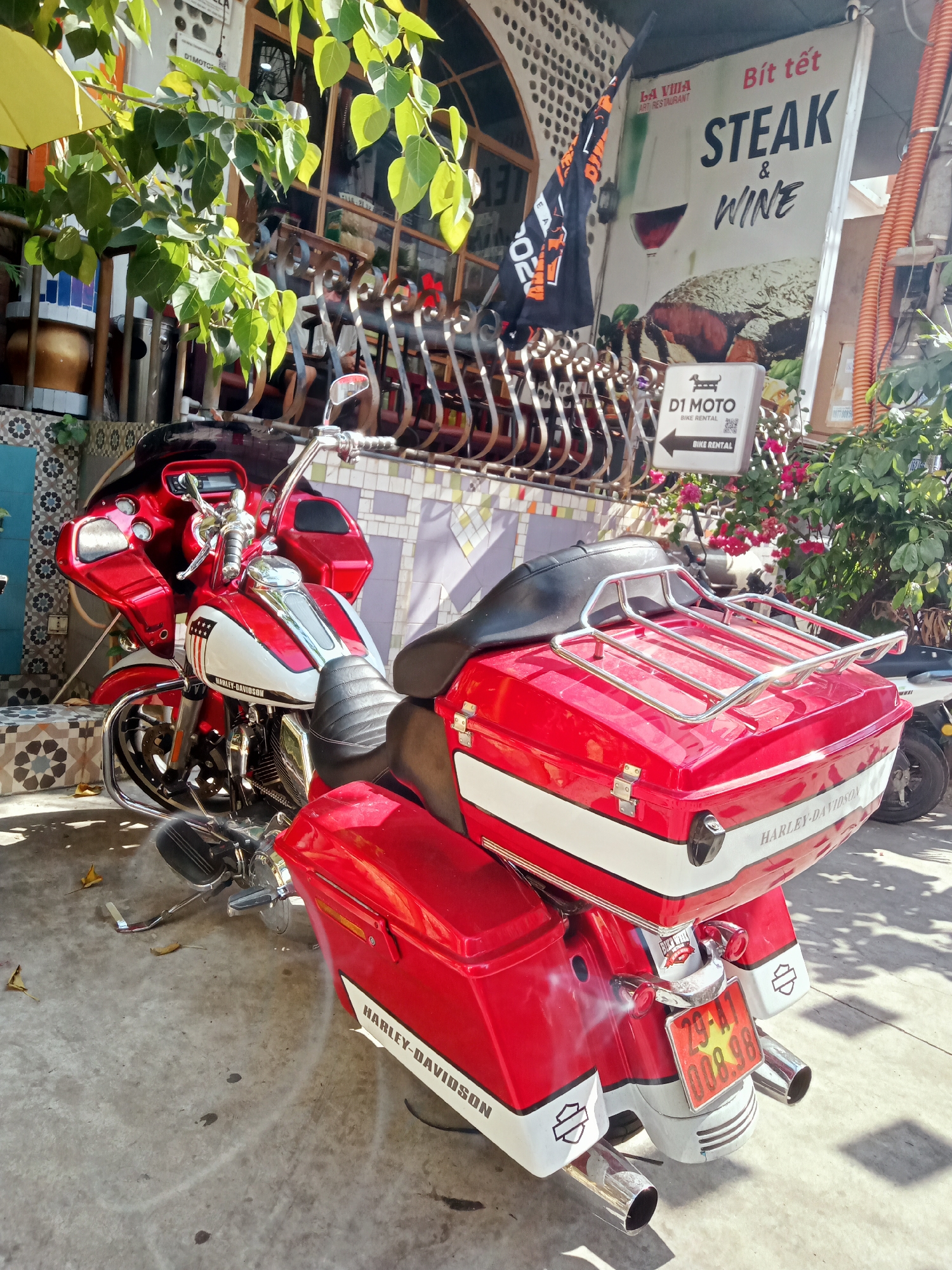 A vibrant red and white Harley-Davidson touring motorcycle is the central subject of this sunlit outdoor scene, parked on a paved area in what appears to be Nha Trang, Vietnam. The motorcycle features a prominent front fairing, ample chrome accents, and matching red saddlebags and a top case, both adorned with white stripes and "HARLEY-DAVIDSON" text. A chrome luggage rack is mounted on the top case. The bike's Vietnamese license plate, "29-A1 008.98", is clearly visible at the rear, below the dual exhaust pipes.
The motorcycle is parked beside a building that houses a restaurant, "LA VILLA ART RESTAURANT," as indicated by a large sign. This sign prominently advertises "Bít tết" (Vietnamese for Steak) and "STEAK & WINE," accompanied by graphics of a wine glass and a steak. Green foliage from trees, with some leaves appearing bright yellow due to direct sunlight, frames the upper parts of the image, casting dappled shadows on the ground.
Beside the restaurant sign, there's a white square sign for "D1 MOTO BIKE RENTAL," featuring a dachshund dog logo and an arrow pointing to the left, suggesting a rental service. A black flag with orange text, partly obscured but showing "D1 MOTOR", hangs near the sign. The overall scene is bright and clear, indicating a sunny day. The style of the architecture and the presence of Vietnamese text strongly suggest a Southeast Asian location, consistent with Nha Trang.