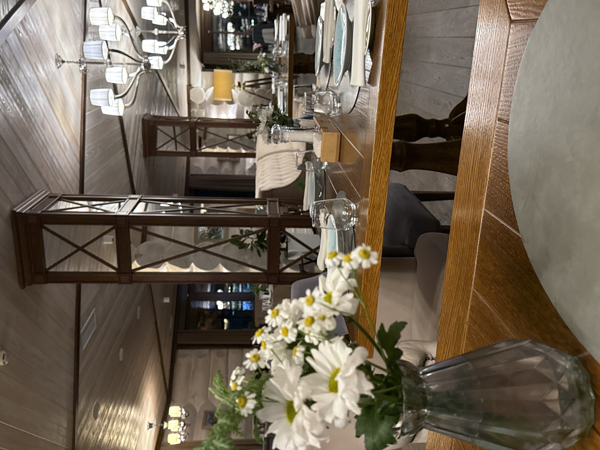 The image depicts the interior of a restaurant in Urlu-Aspak, Russia, characterized by a rustic, wooden aesthetic. In the foreground, a bouquet of white daisies in a textured glass vase sits on a polished wooden table, which is set for a meal with plates, glasses, and cutlery. The table is part of a larger dining area with multiple tables and chairs visible in the background, some occupied by patrons. Chandeliers with multiple bulbs hang from the wood-paneled ceiling, providing warm lighting. The walls are also adorned with wood paneling and decorative elements, contributing to the cozy and traditional atmosphere of the establishment.