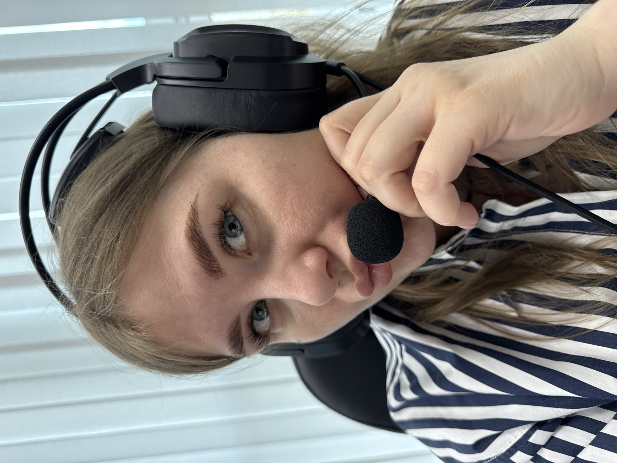 A woman with light skin and blue eyes is wearing black over-ear headphones with a microphone. She is holding the microphone with her right hand, positioning it close to her mouth. She is wearing a dark blue and white striped shirt. The background consists of white blinds. The location of the image is not discernible from the details provided.