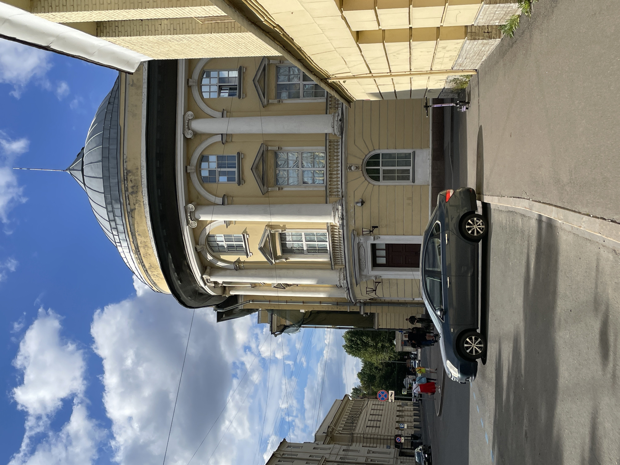 The image shows a street scene in Saint Petersburg, Russia, featuring a distinctive curved building with a dome. The building is painted yellow and features classical architectural elements such as columns and arched windows. To the right of this building, another yellow structure with a brick base is visible. In the foreground, a dark grey sedan is parked on the street. A sidewalk runs alongside the building, and on this sidewalk, a purple electric scooter is standing upright. A few people are visible in the background walking along the street, and some trees line the far side of the street. The sky is blue with scattered white clouds. Traffic signs, including a "no parking" sign, are present on the left side of the street.