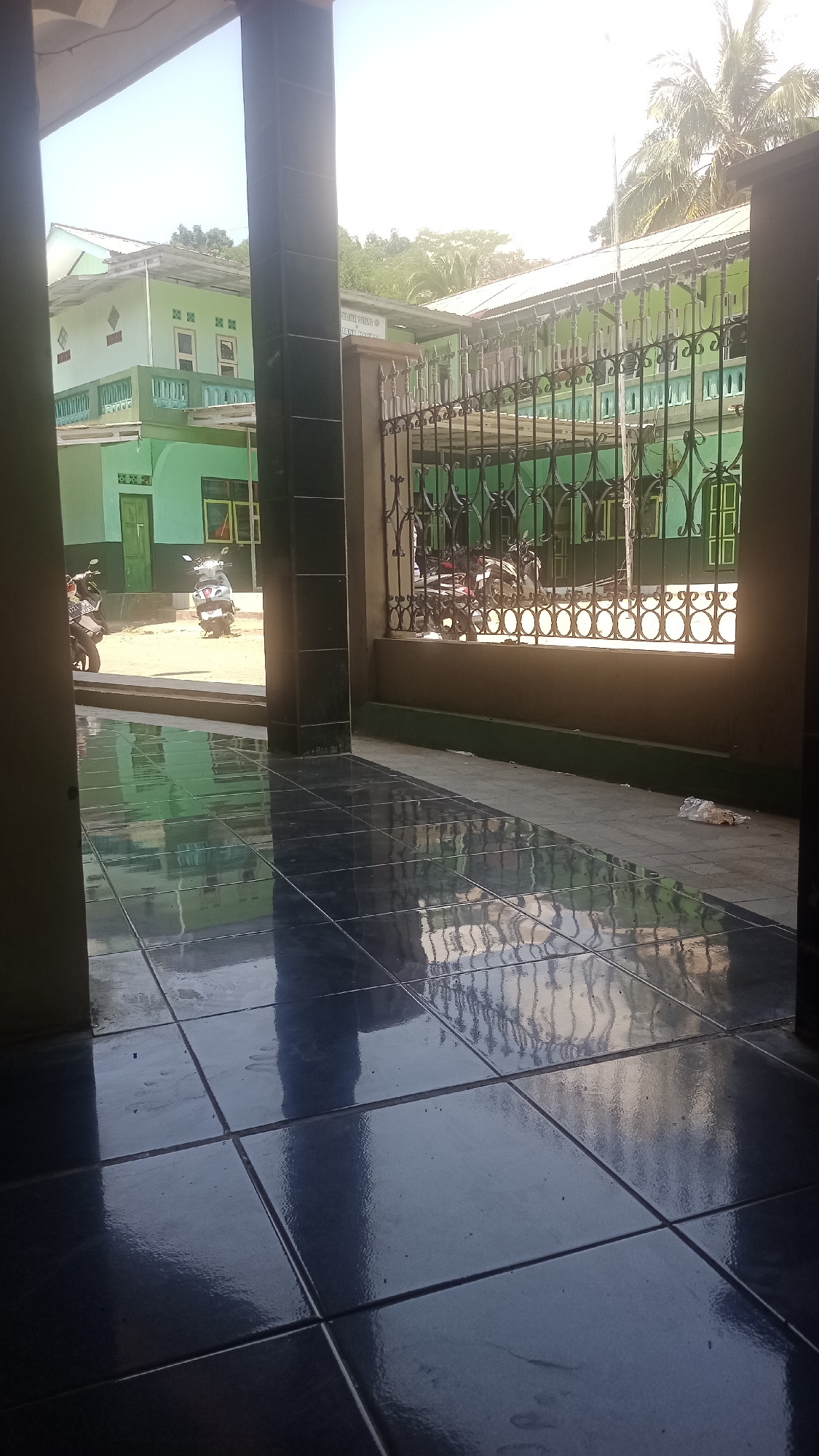 The image depicts an outdoor scene in Karyamandala, Indonesia, featuring a building with green walls and a white roof. In the foreground, a polished tiled floor reflects the surroundings. A dark pillar stands to the left, framing a view of a courtyard with several motorcycles parked. Beyond the courtyard, a fence with decorative metalwork separates the area from a structure also with green walls and a white roof. Trees and palm fronds are visible in the background against a bright sky. The overall impression is that of a public building or complex under daylight.