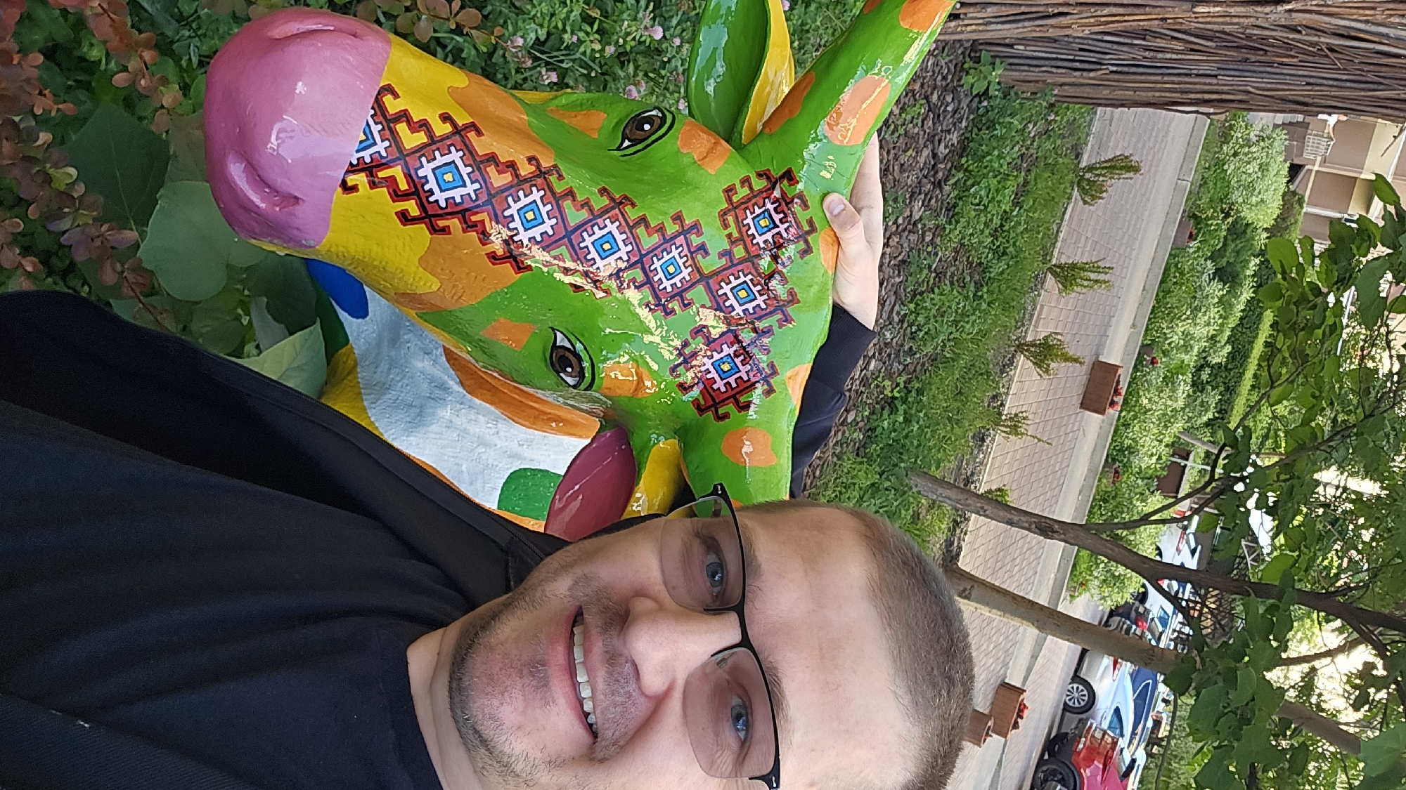 A man with glasses smiles at the camera, taking a selfie. He is standing next to a large, brightly painted sculpture of a cow's head. The cow is predominantly green with colorful geometric patterns. The man is wearing a black t-shirt and jacket. The background shows a tree-lined street with parked cars, suggesting an urban environment. The foliage and pathway indicate an outdoor setting, possibly a park or residential area in Kyiv, Ukraine.