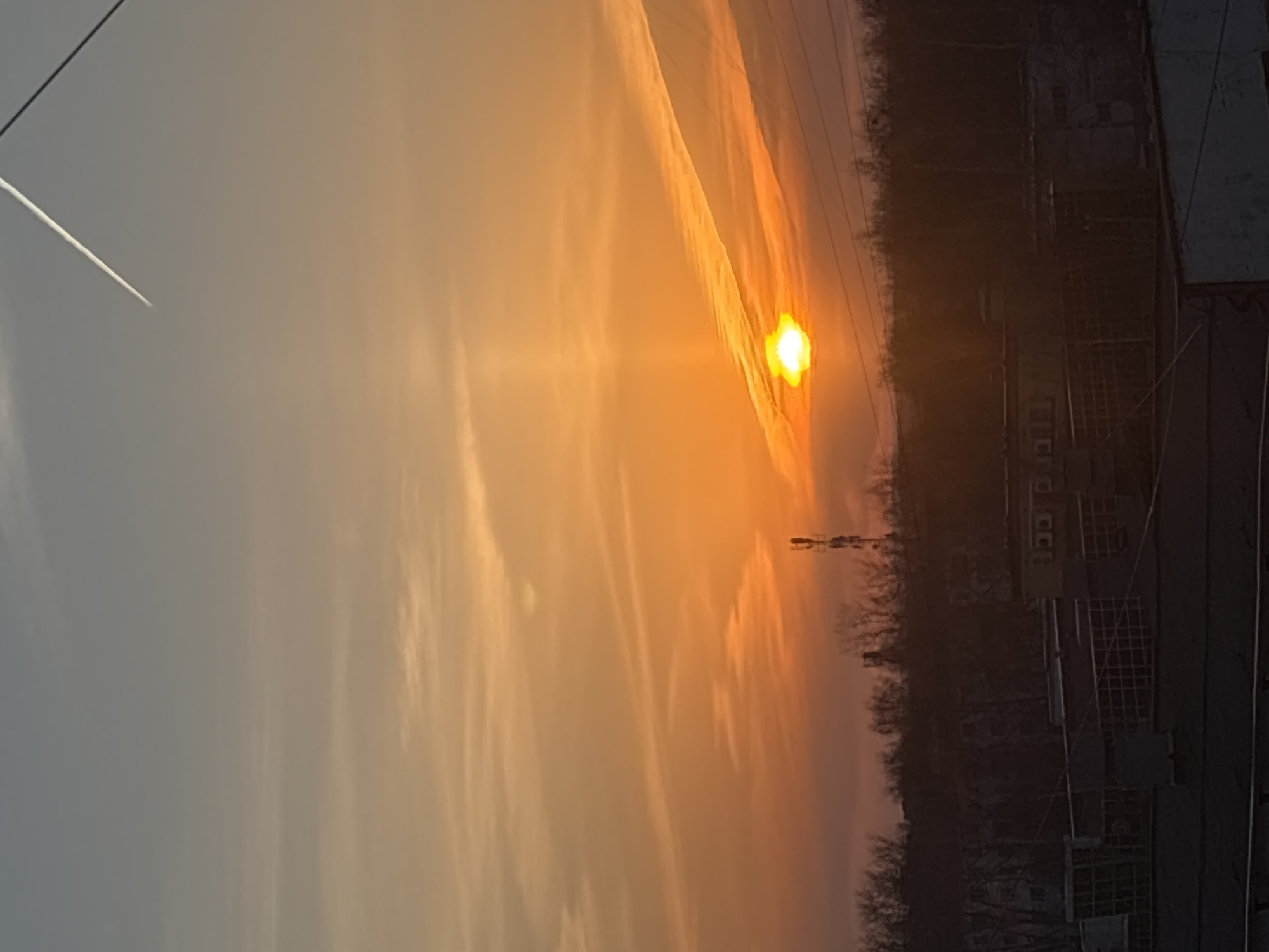 The image shows a sunset in Chaikovsky, Russia. The sun appears as a bright orange orb partially obscured by clouds. Vapor trails of jet pass through the sky. In the foreground, there are buildings and trees silhouetted against the colorful sky. Power lines run across the upper part of the frame.