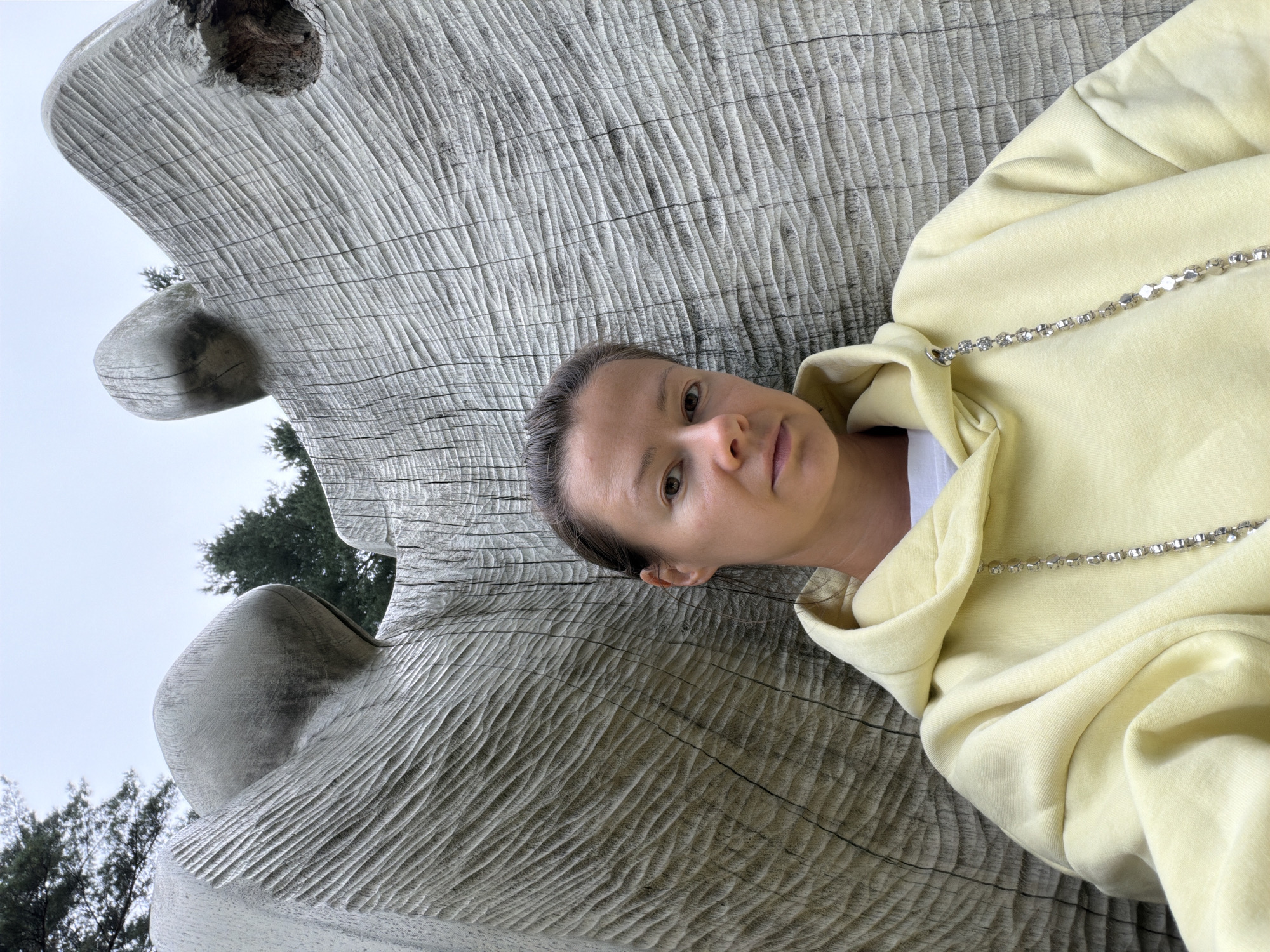 A woman with brown hair, wearing a pale yellow hoodie and a white t-shirt, is photographed from the chest up. She is leaning against a large, rough-hewn wooden sculpture with a textured surface. The sculpture appears to be part of an outdoor installation, with trees visible in the background against a cloudy sky. The scene is likely located in Riga, Latvia.