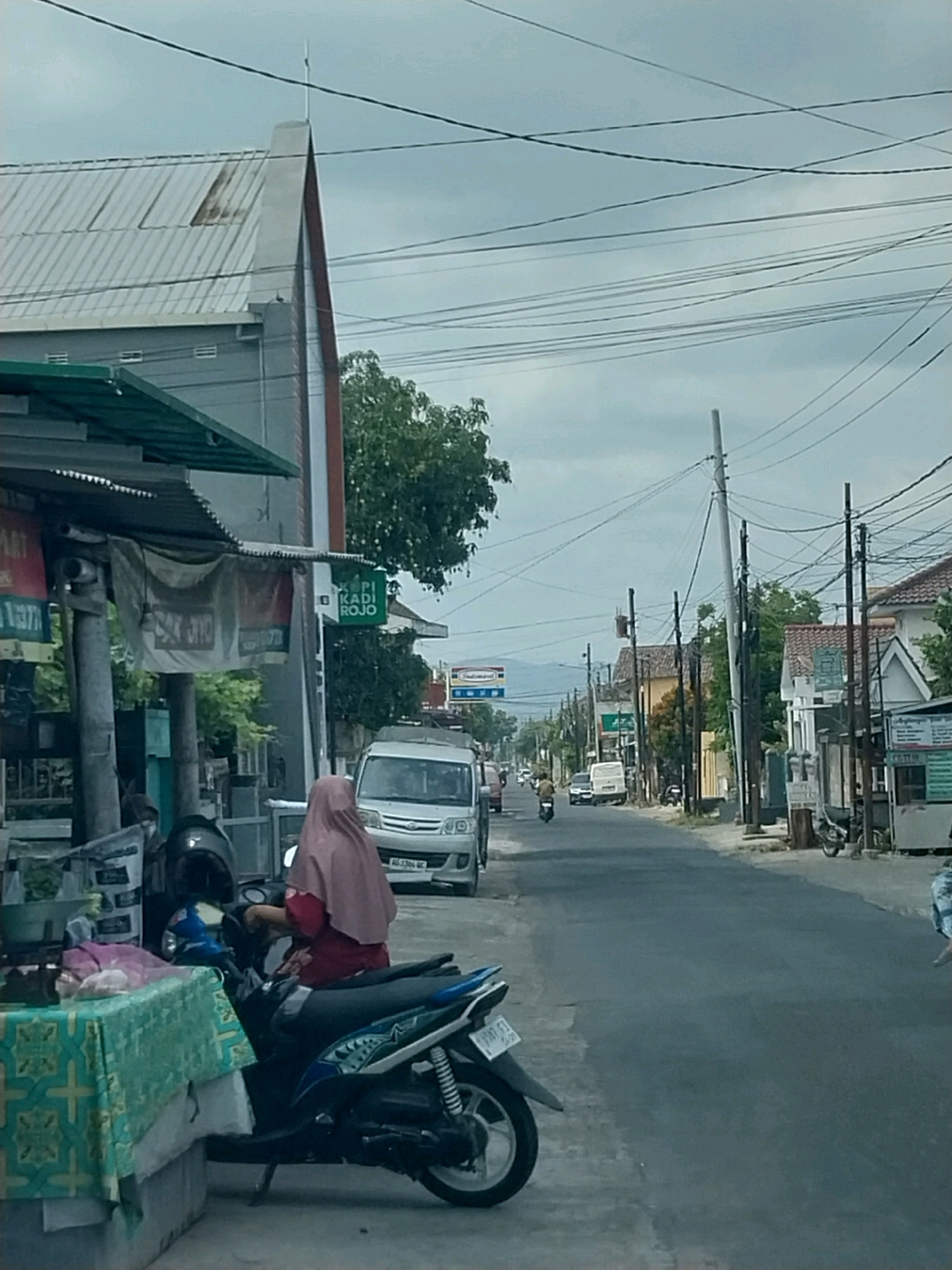 A street view in Purwomartani, Indonesia, shows a paved road with various buildings and trees lining its sides. Overhead, a dense network of electrical wires crisscrosses the sky. In the foreground, a person wearing a pink headscarf and red shirt sits on a dark-colored scooter, parked beside a table covered with a teal and green patterned cloth. To the left, a silver van with a license plate AG-3306 UC is parked on the side of the road. Further down the street, several other vehicles, including motorcycles and cars, are visible, and some individuals are walking or riding their motorbikes. Buildings include structures with metal roofs, some with visible windows and signs, including a green sign with white text that reads "KOPI KADI ROJO." The sky is overcast with a light gray hue.