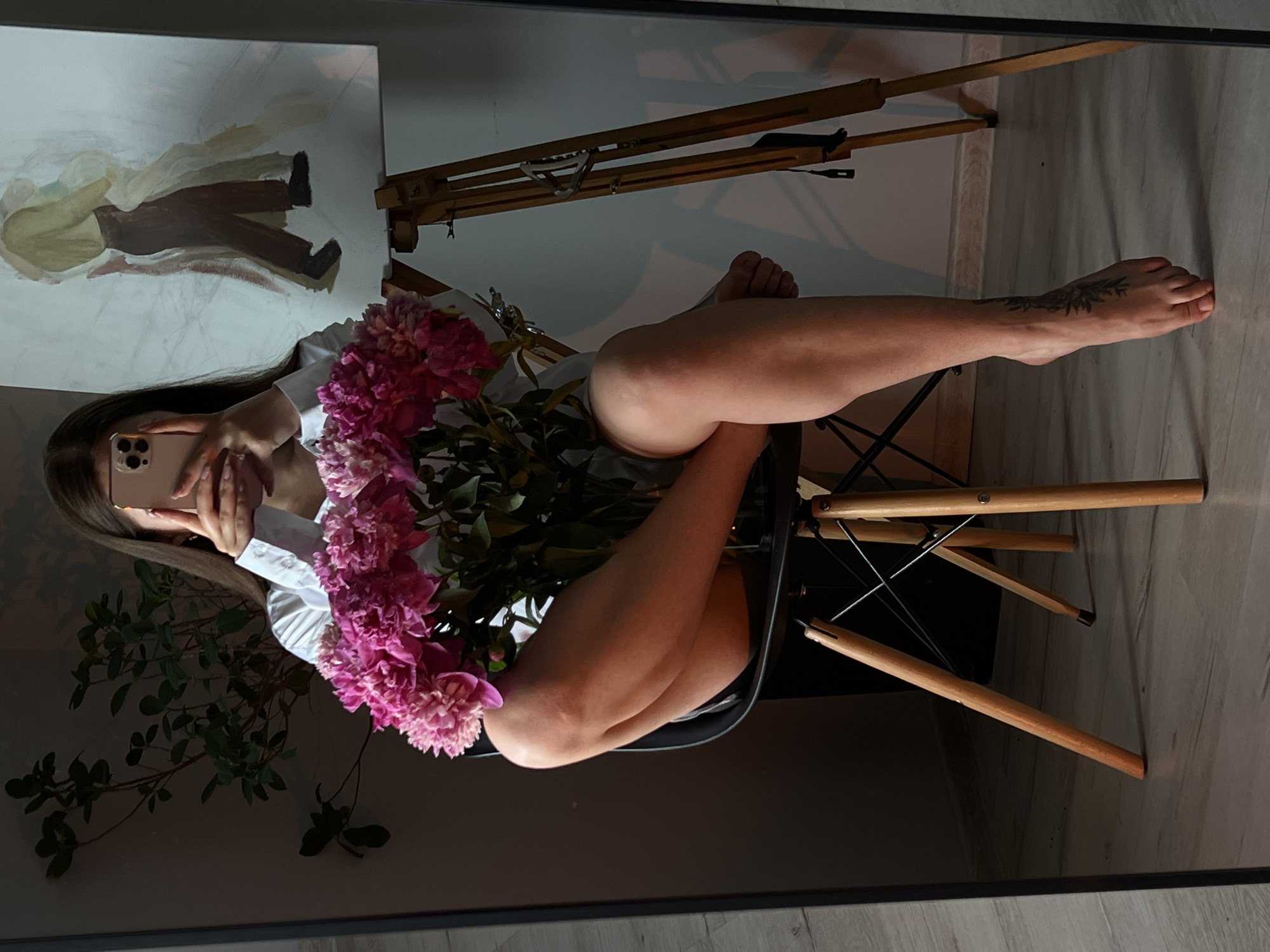 The image shows a woman sitting on a chair in a room, holding a bouquet of pink peonies and a smartphone. She is wearing a white shirt and appears to be taking a selfie in a mirror, as her face is obscured by the phone. To her right, an easel holds a painting of two figures. The room has wooden flooring and a plant in the background. The overall impression is a home interior scene, possibly a studio. The location is identified as Ternopil, Ukraine.