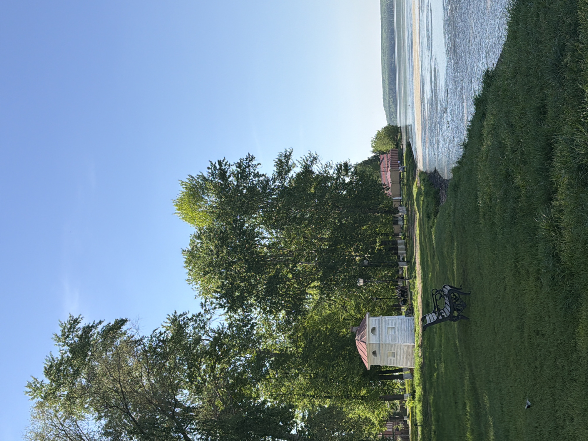 The image shows a park in Votkinsk, Russia. The location is along the shore of the Votkinsk Reservoir, identified by the sandy and muddy banks visible on the right side of the image. A grassy embankment separates the reservoir from the park. A white, two-story building with a red roof stands near a park bench. Numerous trees populate the park, casting shadows on the green lawn. A row of houses and some vegetation are visible along the shoreline in the distance. The sky is clear and blue.