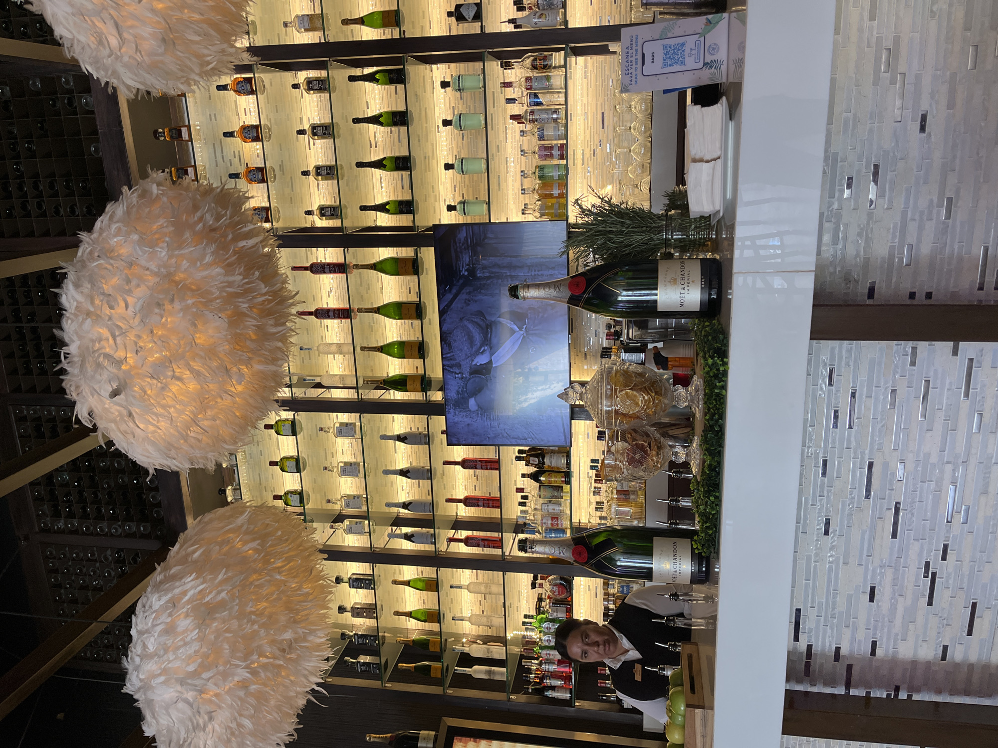 The image depicts the interior of a bar in Playa del Carmen, Mexico. A wall behind the bar is lined with illuminated shelves stocked with various bottles of liquor and wine. A large television screen is centrally located among the shelves. Two large, white, feathered lamps hang from the ceiling. In the foreground, a bartender in a black vest stands behind a bar counter. The bar counter displays bottles of Moët & Chandon champagne, glassware, garnishes, and QR code menus. The bar itself is decorated with white mosaic tiles.