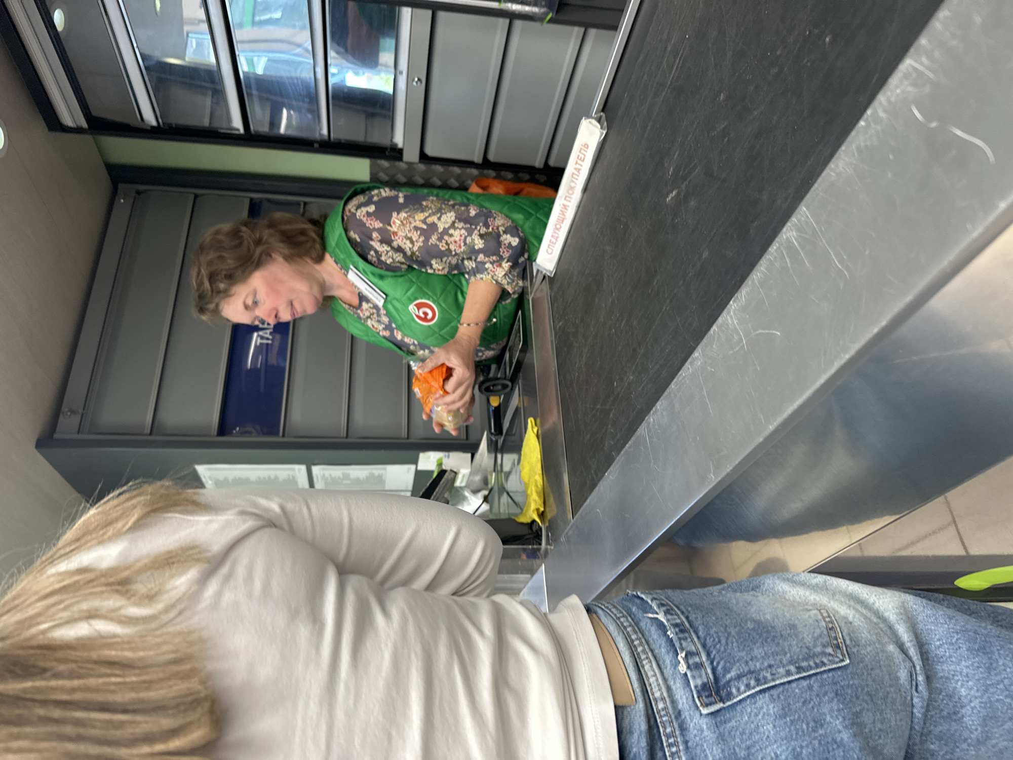A customer stands at a checkout counter in a grocery store in Unknown, Unknown. The customer, wearing a white long-sleeved shirt and blue jeans, faces the counter. A cashier, wearing a green vest and floral print top, is at the checkout station, handling a bag of groceries. The checkout counter has a black surface and a metal bar separating the cashier from the customer. A sign in Russian sits on the metal bar. Shelving and retail displays are visible in the background.