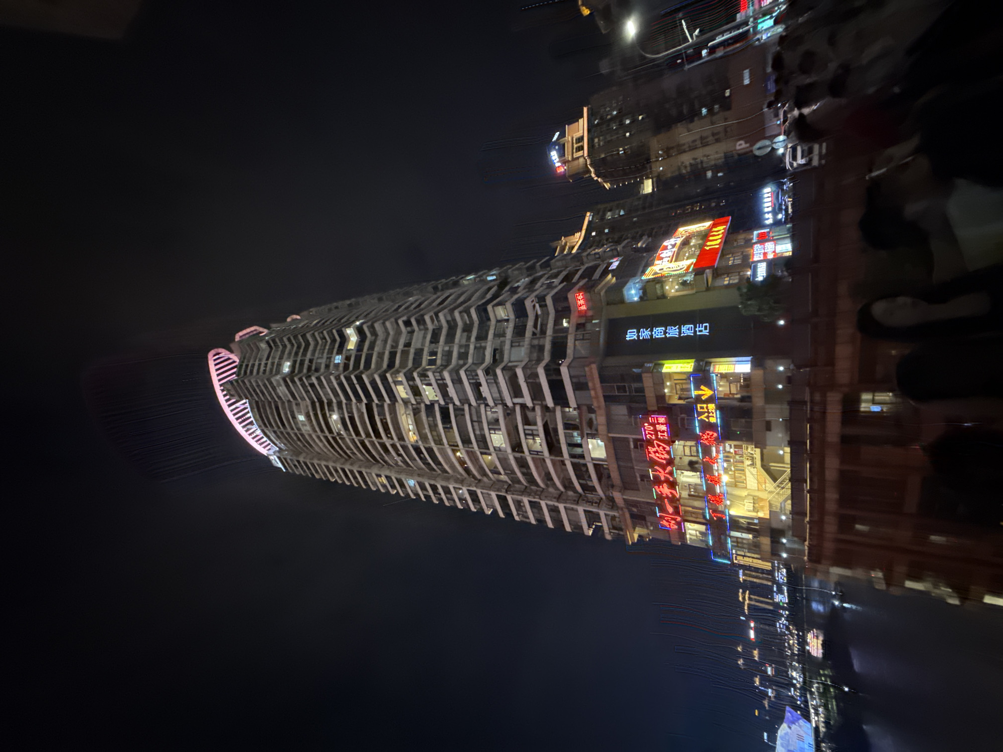 An extreme low-angle shot captures a brightly lit skyscraper at night in Yuzhong District, China. The tower features a distinctive pink and white halo of lights at its apex and is adorned with various neon signs in Chinese characters, predominantly red and yellow, indicating businesses like "Shui Yi Shou Hot Pot" and "Jia Shang Commercial Hotel." The building's curved facade showcases numerous balconies and illuminated windows. In the foreground, the blurry silhouettes of people suggest a crowd observing the scene, possibly from a bridge or elevated walkway, with a body of water and other distant city lights visible below. The overall atmosphere is vibrant and urban, characteristic of a bustling city at night.