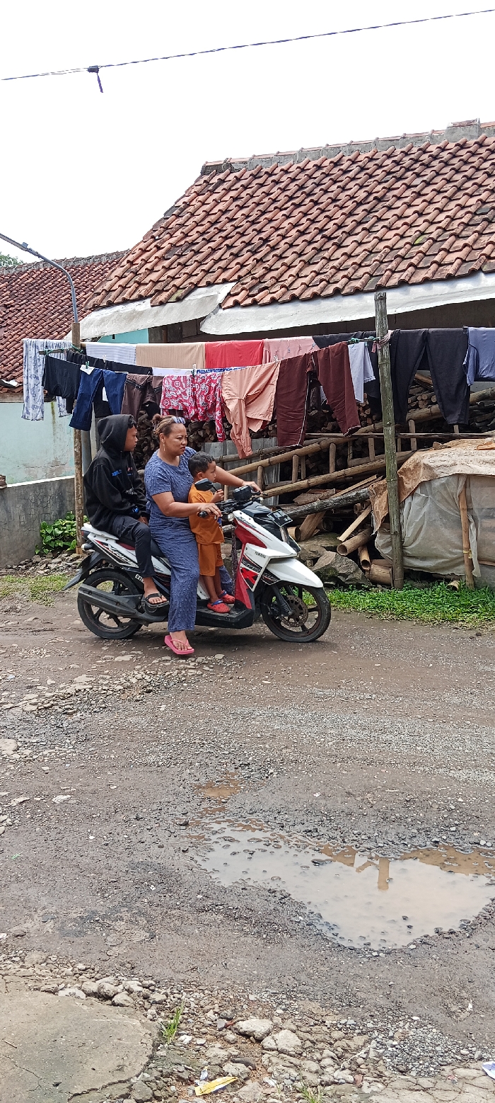 The image depicts a scene in Kutawaringin, Indonesia, with coordinates -6.974, 107.519. Three individuals, including a woman and two children, are situated on a white Yamaha motorcycle. The woman is wearing a brown shirt and blue pants, while the children are wearing orange shirts. The motorcycle is parked on a dirt road in front of a residential building with a red tiled roof. Several pieces of laundry are hanging on a clothesline in front of the building. The road is rough and uneven, with a small puddle of water reflecting the surrounding environment. The scene suggests a typical residential area in a developing country.