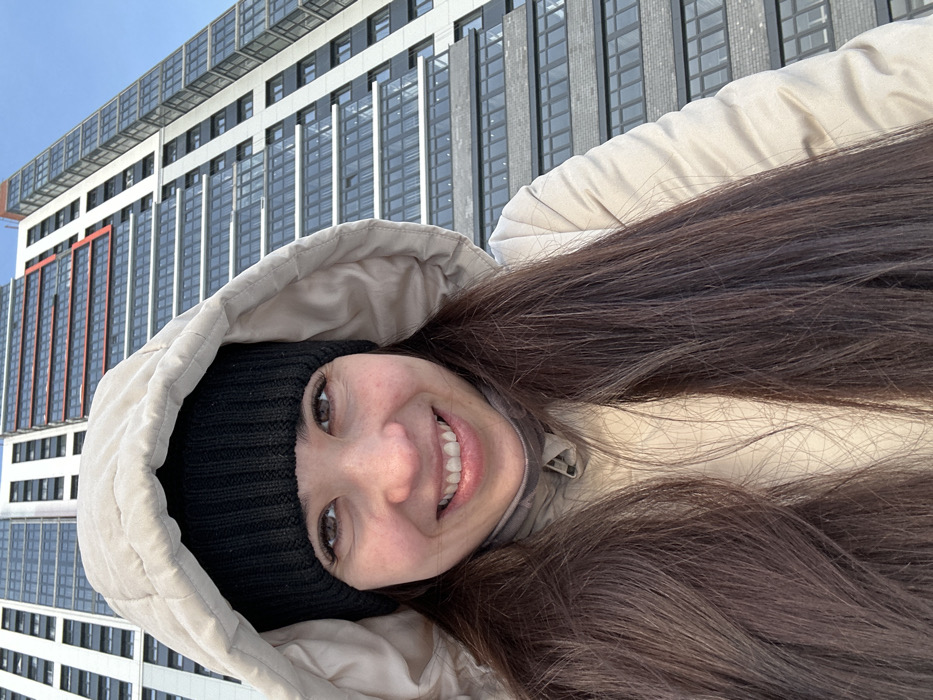 A low-angle, close-up photograph captures an adult female smiling directly at the viewer in Dikaya Gar', Russia. She possesses long, straight, dark brown hair, dark eyes, and is attired in a black knitted beanie and a beige quilted hooded jacket, with the hood up and the jacket zipped to her chin. In the background, under a clear light blue sky, stands a modern high-rise building. This structure features a facade composed of multiple horizontal bands of dark glass windows interspersed with light-colored panels. Distinct orange-red architectural elements are visible on the upper levels of the building.
