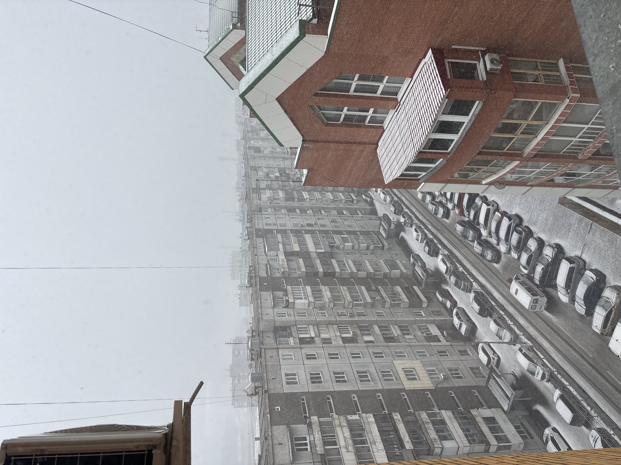 A high-angle, eye-level shot shows a cityscape in Krasnoyarsk, Russia during snowy weather. Predominantly gray, multi-story apartment buildings with numerous windows and external air conditioning units stretch into the distance. Cars parked along a street run adjacent to the buildings. A red brick building with white-framed windows and a green-trimmed roof appears in the foreground. A cable line stretches across the sky. The scene is overcast and the snow is falling steadily.