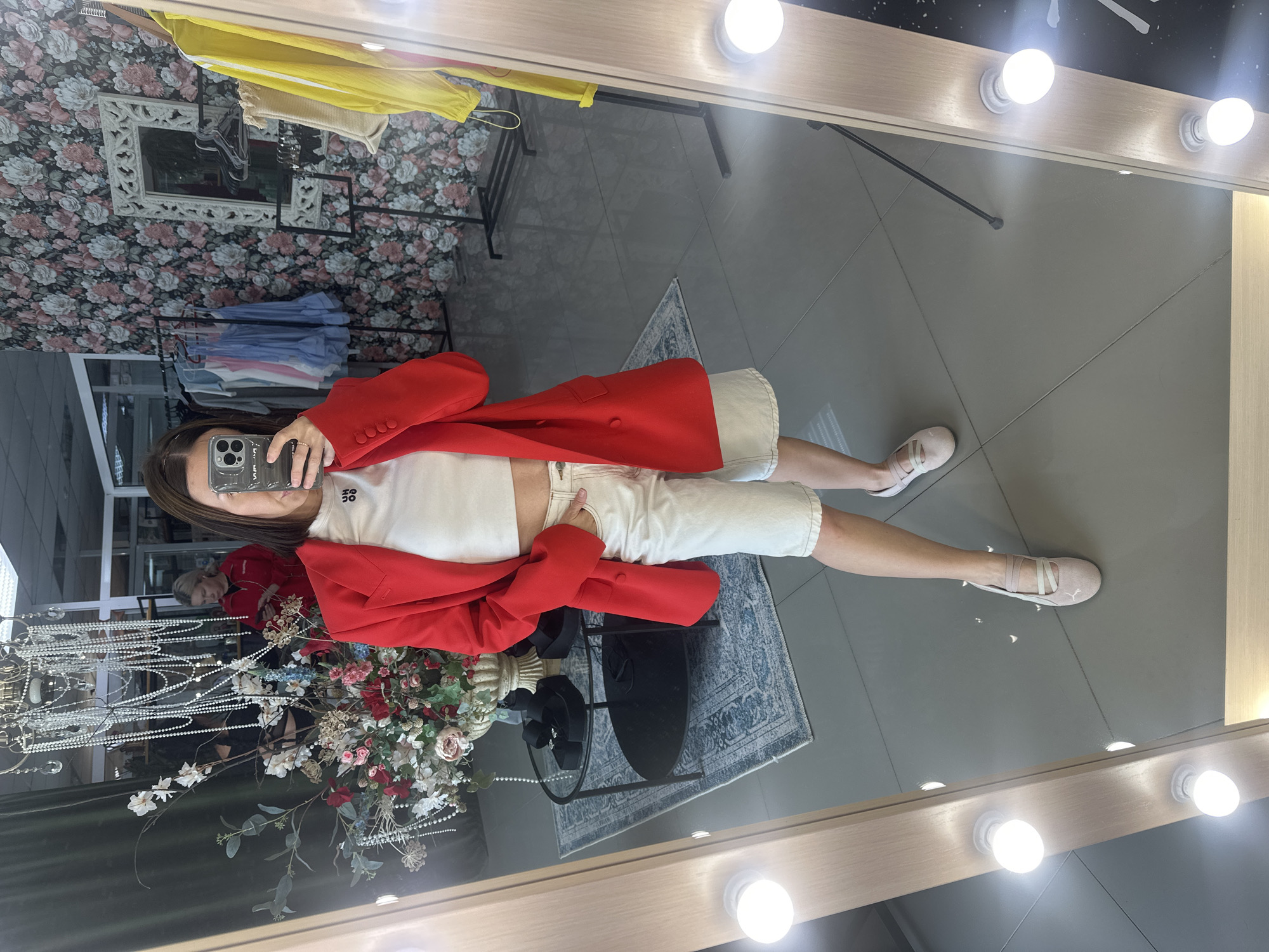 A woman is taking a mirror selfie in what appears to be a retail store. She is wearing a red blazer over a white crop top and white shorts. She has her right hand in her pocket. In the background, there are clothes on racks, a chandelier, and a floral wallpaper. A blurred figure in a red jacket is visible in the reflection of a mirror to the left. The floor is tiled in gray. The mirror she is taking the selfie in has lights around its frame. The store is located in Votkinsk, Russia.