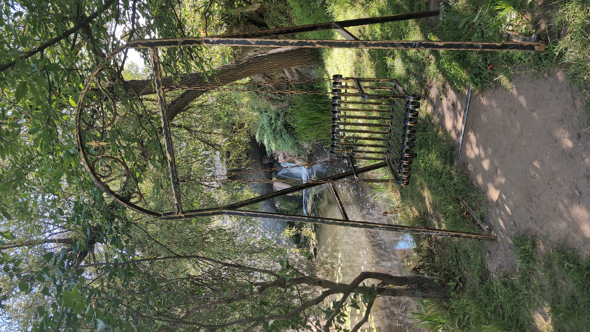 An outdoor scene depicts a metal swing set positioned on a dirt path bordered by grass. The swing set has an arched top with decorative metalwork and a seat made of metal bars. To the left of the swing, a body of water with lily pads is visible, bordered by a rocky embankment and a small waterfall. Lush green trees with dense foliage surround the area, casting shadows on the ground. The overall environment suggests a park or a natural setting. There are no people present in the image. The location is identified as being in Radomyshl, Ukraine.