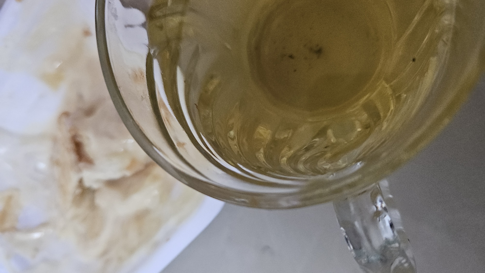A top-down shot depicts a glass cup filled with a yellowish liquid, possibly tea, placed on a white surface. To the left, a blurry, white plate holds food with a mottled tan and white appearance. The glass cup has a clear, ribbed design. The scene is in Cam Đức, Vietnam.