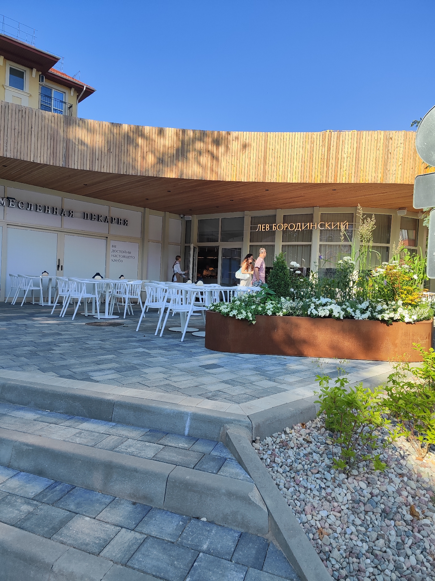 This is a medium shot, taken outdoors, of a modern bakery and cafe with outdoor seating. The building features a curved wooden canopy over a facade of large windows. To the left, above the entrance, is text that reads "МЕСЛЕННАЯ ПЕКАРНЯ" which translates to "Maslenitsa Bakery". To the right, over another section of windows, is text reading "ЛЕВ БОРОДИНСКИЙ". Inside the cafe, several tables and chairs are visible. In front of the building, white tables and chairs are set up on a paved area. A large, rusty metal planter filled with white flowers and greenery sits in front of the right side of the cafe.
Two people are visible walking near the entrance to the cafe. One person, wearing a light-colored shirt and shorts, is walking away from the camera, while another person, wearing a light-colored dress and carrying a bag, is walking towards the camera. A person in a white uniform is seen working inside the cafe. The sun is shining, casting shadows from the wooden canopy onto the building and pavement, suggesting it is daytime. The sky is clear and blue. The scene conveys a pleasant, relaxed atmosphere of an outdoor cafe on a sunny day. The paving stones and the design of the building suggest a contemporary urban setting.