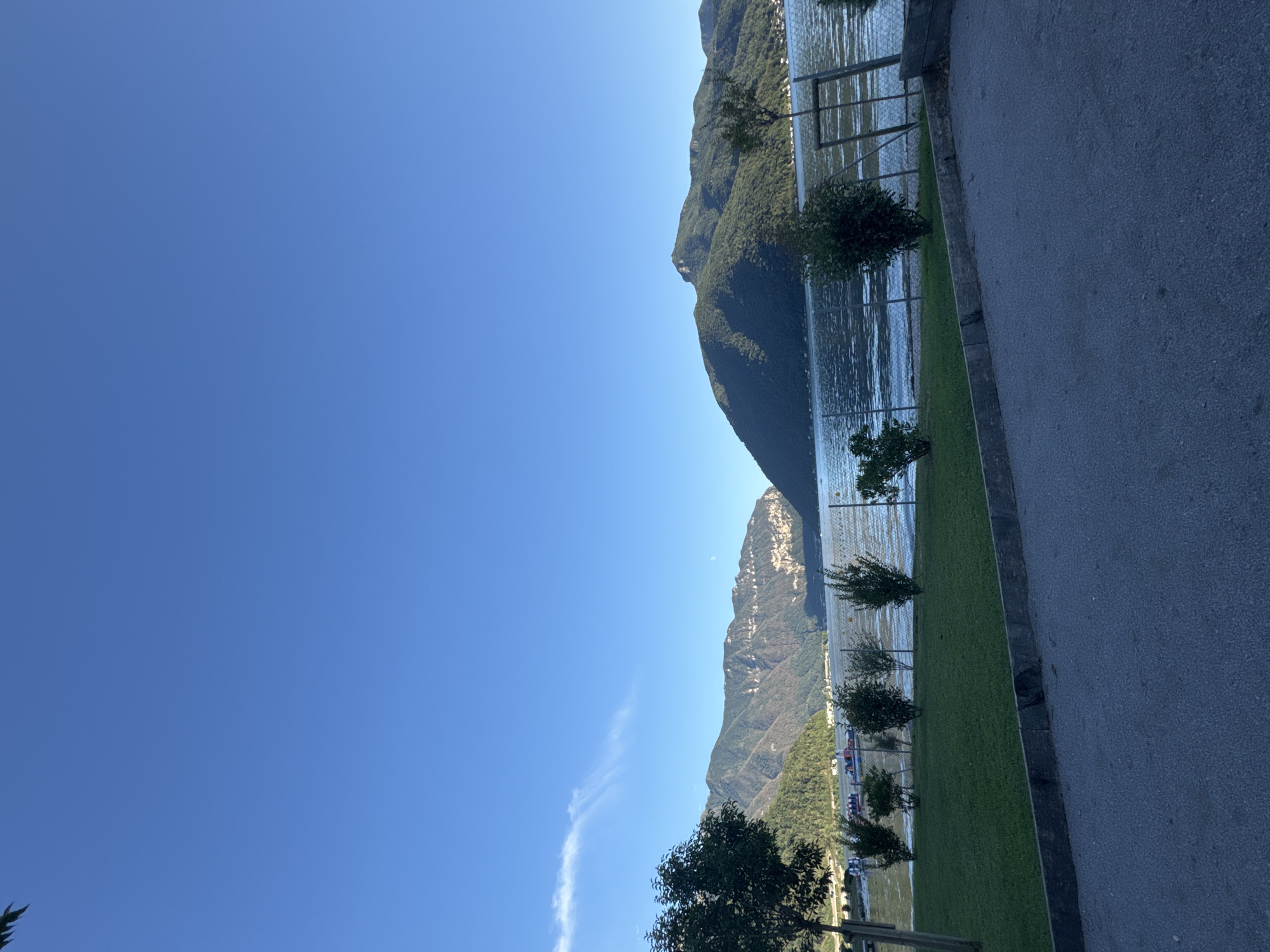 The image captures a serene view of Lake Lugano in Melide, Switzerland, under a clear blue sky. In the foreground, a paved surface transitions to a manicured lawn dotted with small, green shrubs. A metal fence separates the grassy area from the lake. Several small, colorful boats are visible on the water's edge, some of which appear to be paddle boats. Across the lake, lush green mountains rise, their slopes covered in dense vegetation. The distant mountain range exhibits a rugged, layered topography. The sunlight illuminates the scene, casting soft shadows. The atmosphere is peaceful and tranquil, suggesting a pleasant day by the lake.