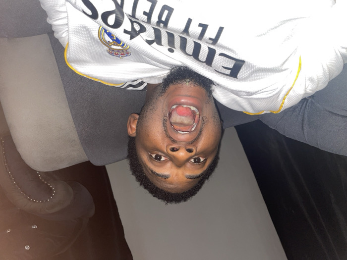 The image depicts an adult male situated indoors, stated to be in the city of Ube, Nigeria. The subject, with dark skin, short dark hair, and a goatee beard, is positioned centrally and looking directly at the camera. His mouth is open wide, revealing his tongue and the oral cavity. He is wearing a white short-sleeved shirt featuring the 'Emirates FLY BETR' sponsor logo and the Real Madrid club crest, with yellow detailing on the collar or shoulder seam. The individual is seated on a dark grey upholstered chair or sofa. In the background to the right, a section of another dark upholstered furniture piece, adorned with studded detailing, is partially visible. To the left background, a dark, indistinct surface, possibly a curtain or wall, is present. The scene is illuminated, highlighting the subject's face against a generally dim background.