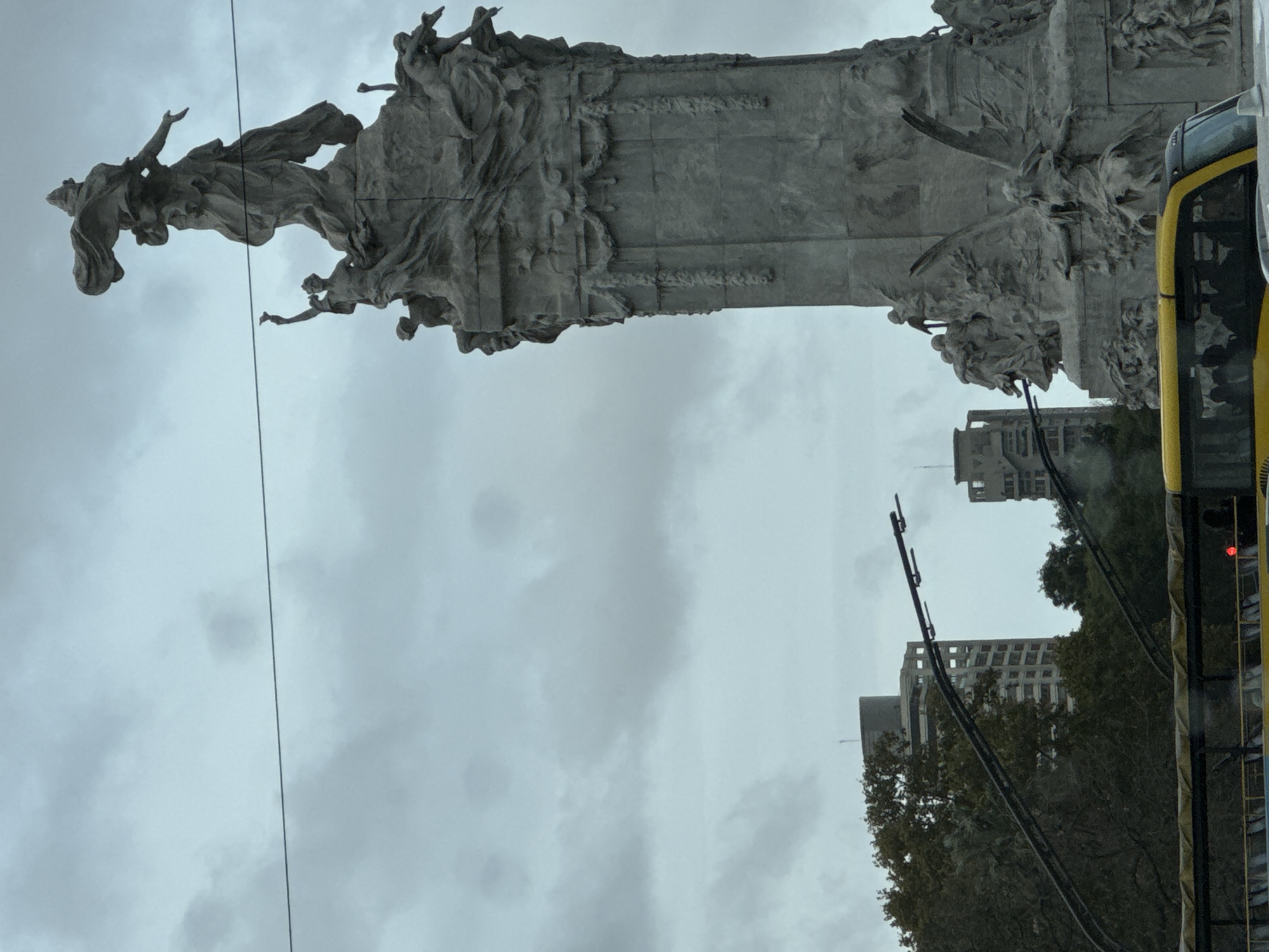 The image shows a partial view of the Monument to the Carta Magna and Four Regions of Argentina, located in the city of Buenos Aires, Argentina. The monument features a sculptural group with allegorical figures on top of a tall pedestal. In the background, buildings can be seen under a cloudy sky. The right side of the image shows the side of a yellow bus.