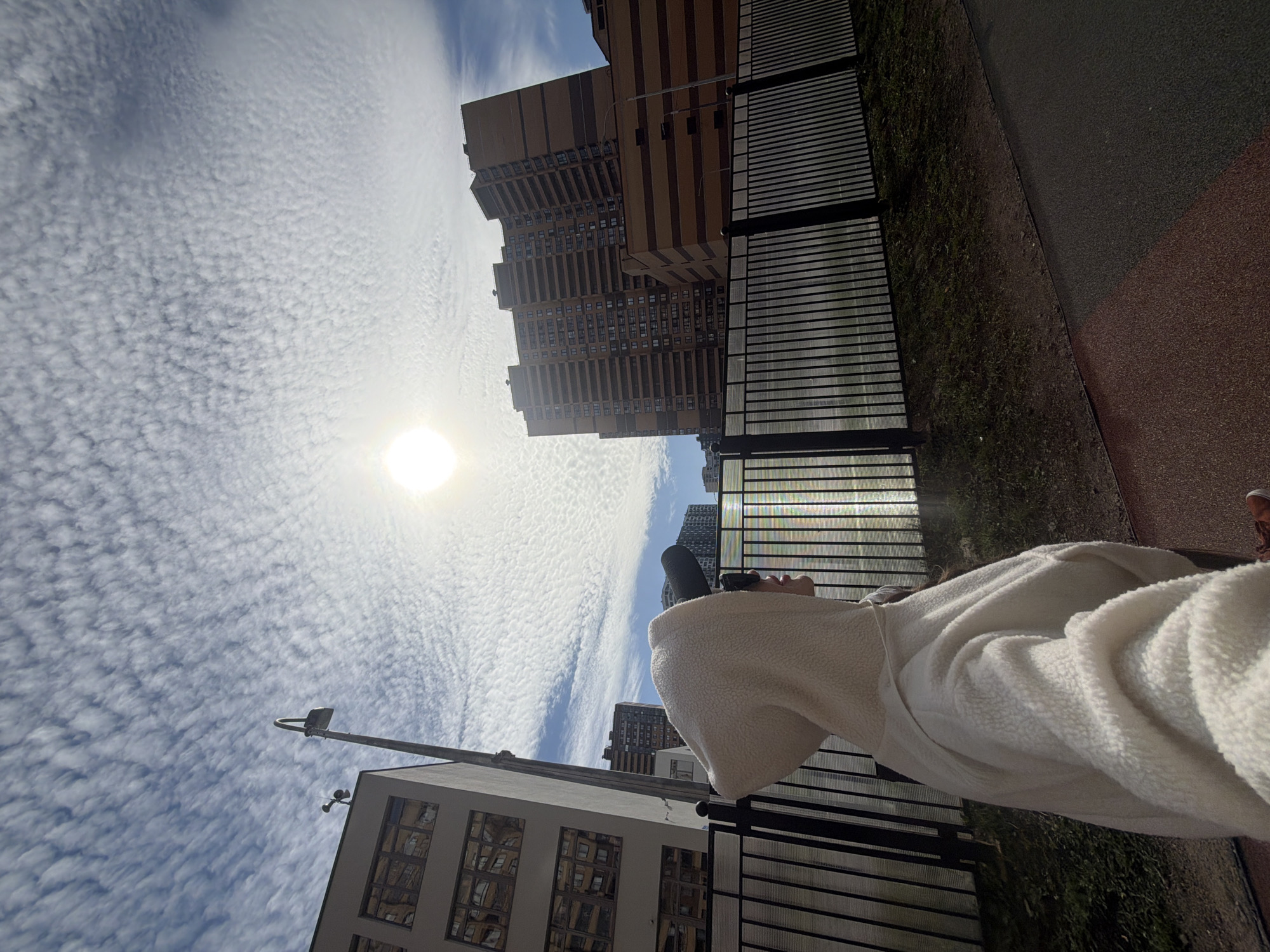 A ground-level, oblique photograph depicts a person, partially visible from the shoulders down, wearing a white, textured jacket, standing near a black, horizontal-bar fence. The sun is high in a sky filled with scattered clouds. In the background are urban buildings, one a tall, multi-story structure with a light brown façade and dark brown horizontal stripes. Another building is lower, with a white exterior and multiple windows. A streetlight is visible in front of the white building. Adjacent to the fence is a narrow strip of green vegetation and a red-and-black surface. The photo's orientation is unconventional, taken at an angle that shows both the person and the surrounding cityscape. The location is likely in the city of Saint Petersburg, Russia.