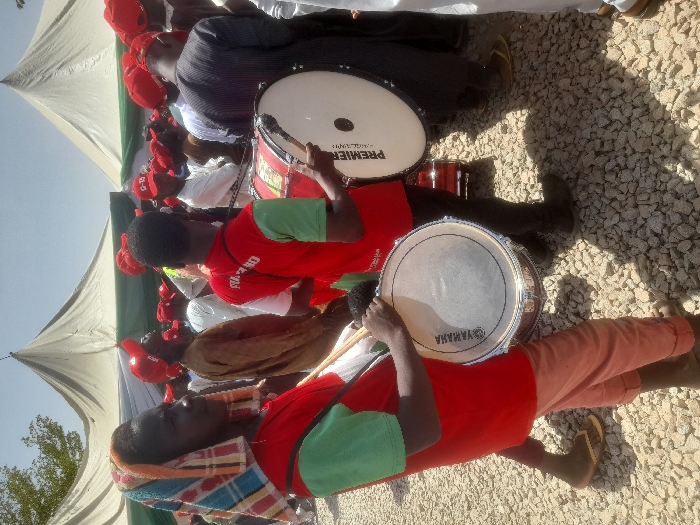 The image shows a scene at a political rally in Gusau, Nigeria (latitude: 12.156, longitude: 6.709). The scene is set outdoors under two large white tents. A group of people in red shirts, some wearing red caps, are gathered in front of the tents. There are at least two drummers, one playing a Yamaha snare drum, the other playing a Premier drum. There is a large crowd of people standing and sitting in the background. The ground is covered with loose gravel, and the sky is partly cloudy. The overall mood of the scene is one of excitement and anticipation.