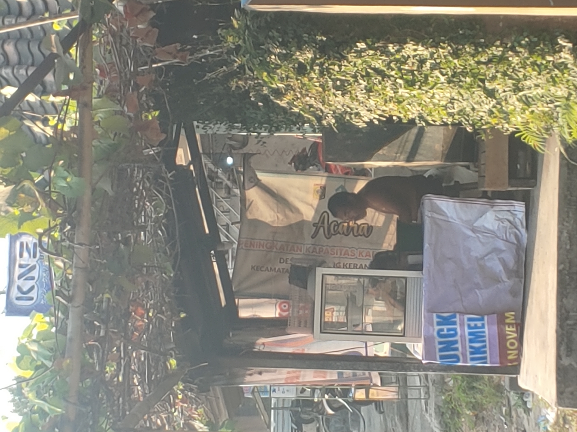 A man is sitting at a stall in Kembang Kerang, Indonesia, with a glass display case in front of him. The stall is covered by an overhead structure made of bamboo and vines, with a tiled roof. In the background, there are banners and signs, one of which appears to be for "KNZ" with some text underneath. The man is wearing a brown shirt and appears to be looking down. There is a white plastic bag with blue lettering that reads "UNGKIKMEL NOVEMBER" in front of the stall. Green foliage is visible on the right side of the image, and in the background, there is a glimpse of a motorcycle. The overall scene suggests a street-side vendor or small shop.