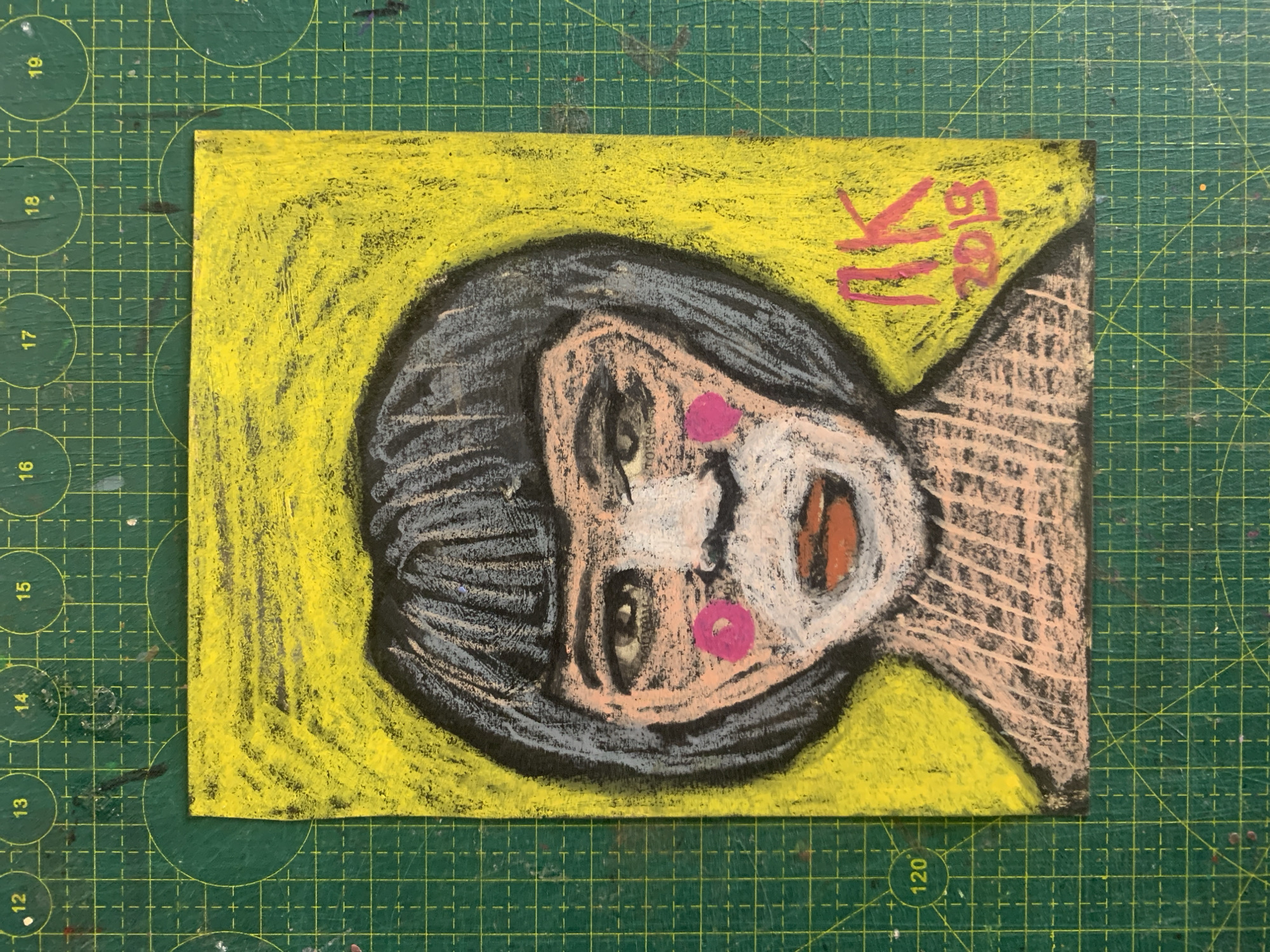 The image is a close-up, eye-level photograph of a pastel drawing of a person's head and shoulders. The drawing is on a yellow background with a black outline. The person has dark hair, grey and black streaks in the hair, light skin with pink circles on the cheeks, white around the nose and mouth, and orange lips. There are also black eyebrows and greyish-blue shadows around the eyes. The person is wearing a light pink garment with black stripes. In the bottom right corner of the drawing, there is red text that appears to read "AK 2018". The drawing is placed on a green cutting mat with grid lines and circular markings indicating measurements in centimeters. The numbers 12, 13, 14, 15, 16, 17, 18, 19, and 120 are visible on the cutting mat. There is no indication of Sergeevo, Russia, in the image or the surrounding environment.