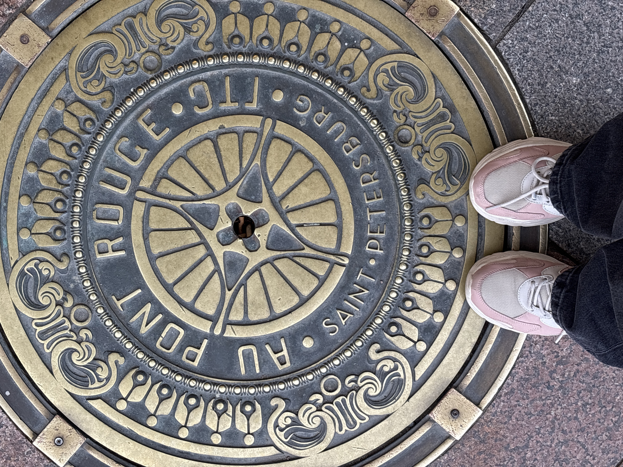A top-down, full shot captures a bronze-colored manhole cover, positioned on a sidewalk, with a pair of pink and white sneakers visible in the upper right quadrant. The manhole cover features a circular design with ornate, baroque-style embellishments along the outer rim. Inscribed in a circular arrangement around the central hub are the words "ROUGE • PONT • AU • SAINT • PETERSBURG • ITC". The hub showcases a stylized compass rose design. The sneakers belong to a person wearing black jeans. The sidewalk surface is composed of grey stone tiles. The scene is most likely located in Saint Petersburg, Russia, given the inclusion of "Saint Petersburg" on the manhole cover.