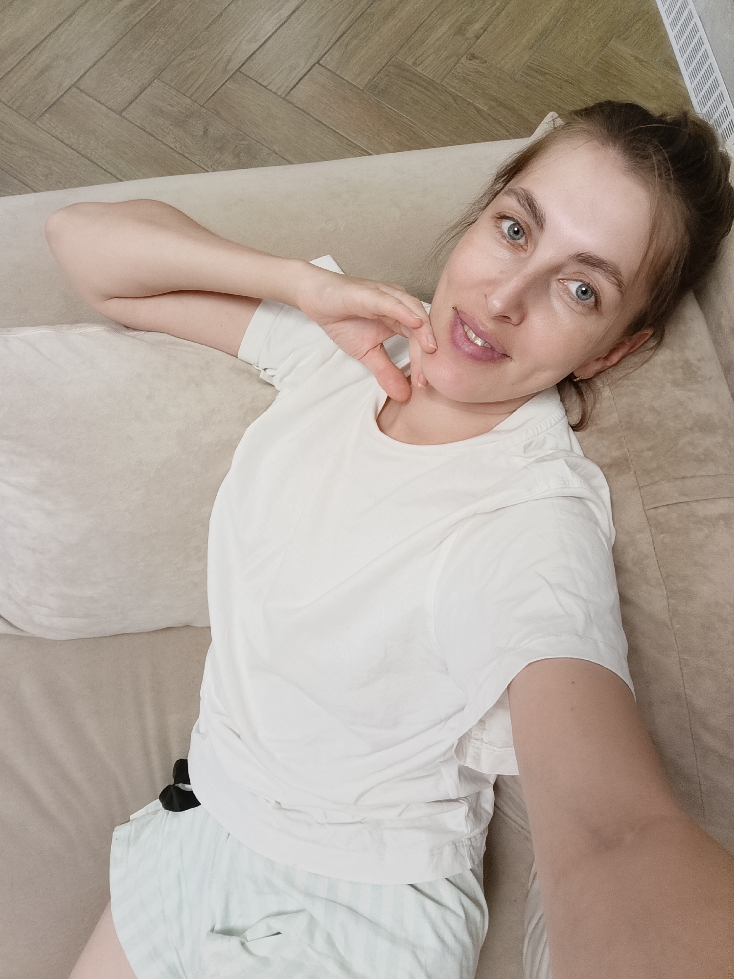 A woman with light brown hair pulled up in a bun, blue eyes, and a subtle smile is lying on her side on a light brown sofa. She is wearing a white t-shirt and light green striped shorts. Her left arm is resting behind her head, and her right arm is bent, with her hand touching her chin. The background shows a wooden floor with a herringbone pattern and a white baseboard. The scene is indoors, likely a residential setting, in the city of Chaikovsky, Russia.