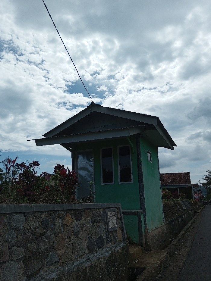 The scene depicts an outdoor environment in Ariyalai, Sri Lanka, under a sky characterized by scattered cumulus clouds and patches of blue. The primary subject is a small, single-story structure, approximately eight to ten feet in height, painted a light green color. It features a dark-colored gable roof with a corrugated metal overhang. An electrical wire extends diagonally upward from the apex of the roof towards the upper left. The building has two rectangular, dark-paned windows on its front-facing wall, and a smaller, similar window on its right-side wall. To the left of the prominent windows, a reflective, possibly metallic, panel or door is visible.
In the foreground, a low, robust stone wall, constructed from irregularly shaped, multi-colored rocks, spans the lower portion of the image. A flat concrete coping caps the wall, and a small, rectangular plaque is affixed to its lower right section. Behind this wall, to the left of the green building, a dense cluster of reddish-purple leafy plants and other green foliage is present.
Immediately in front of the stone wall and running parallel to it is a narrow concrete drainage ditch. Adjacent to the ditch, a paved road extends into the background, curving slightly to the right. In the distant midground, additional residential structures with reddish-brown tiled roofs are partially visible. No individuals, actions, or direct interactions are evident within the frame. The overall lighting is soft and diffused, consistent with an overcast or partly cloudy day.