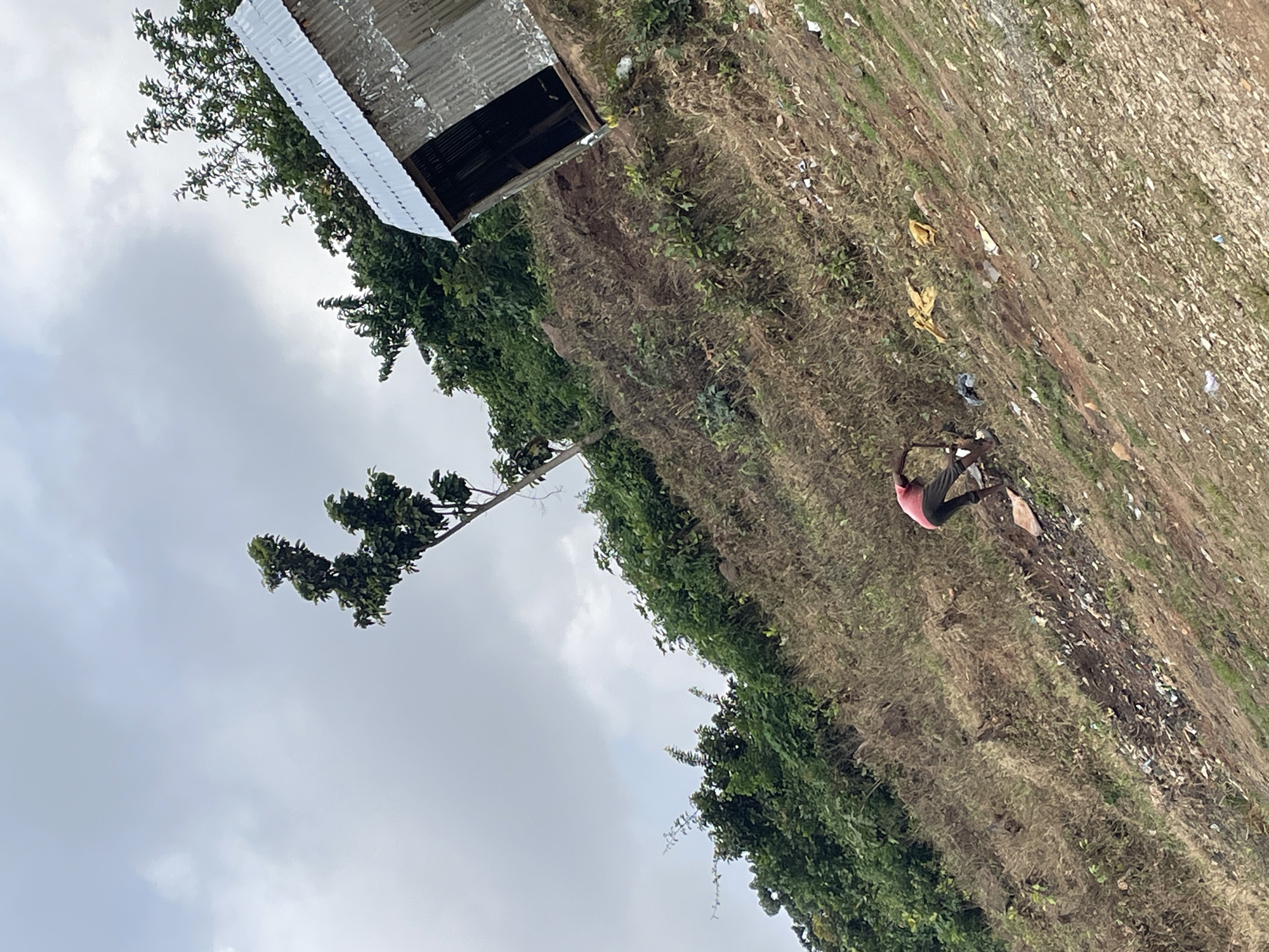 In Kurunduma, Nigeria, a person in a pink shirt and dark pants is working on a steep, overgrown slope. The person is crouched down, appearing to dig with a tool. Debris, including plastic bags and other trash, is scattered across the hillside. In the background, a small, dilapidated structure made of corrugated metal stands on the upper part of the slope. Lush green trees and vegetation surround the area, and the sky is overcast.