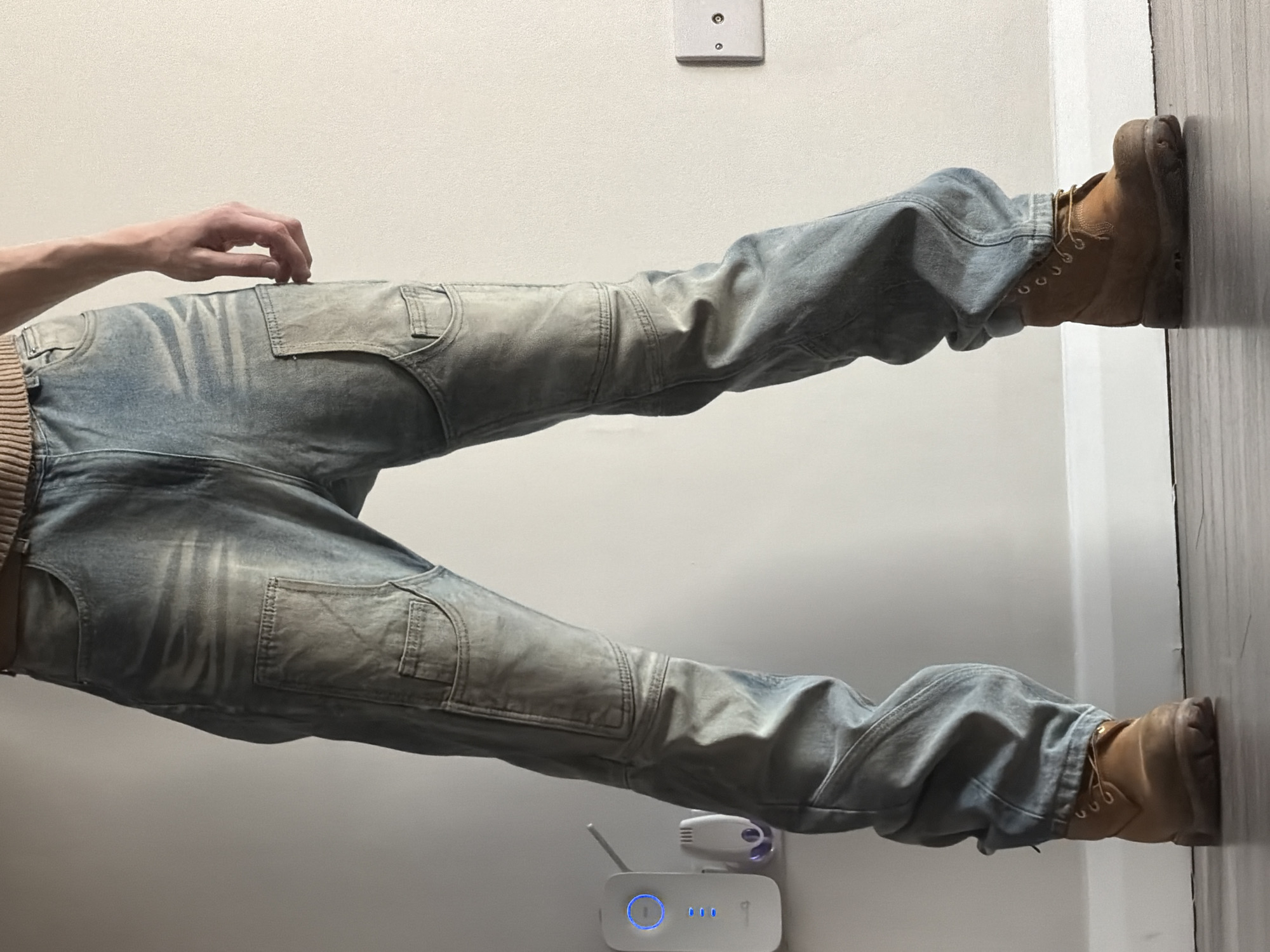 A person is wearing distressed, light blue jeans and tan work boots. Their feet are pressed against a white wall, possibly for balance or support. The wall is adjacent to a light-colored wall with an electrical outlet near the top. Near the lower legs is a white electronic device with a small antenna and blue lights.