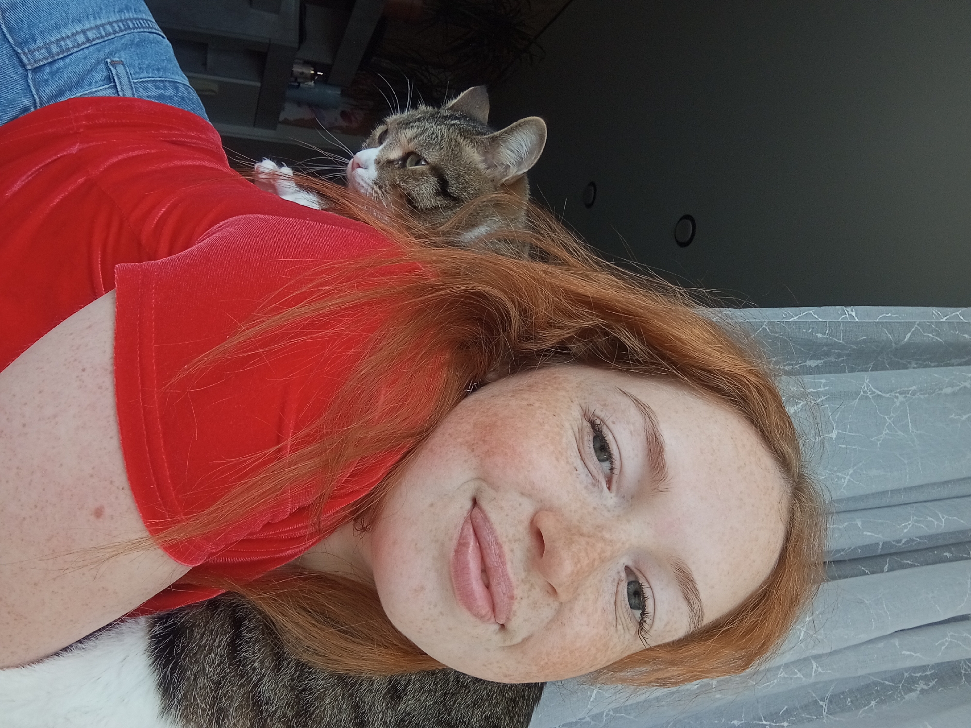 This is a horizontally oriented close-up photograph featuring a young woman and a domestic cat in an indoor setting. The woman, with fair skin and numerous freckles, has long, reddish-auburn hair. She is smiling directly at the camera, her lips slightly parted, revealing light pink lips. Her eyes are light-colored, possibly grey or blue. She is wearing a red top with a velvet or corduroy-like texture, and blue denim pants are partially visible in the upper left corner.
A tabby cat, with striped brown, grey, and white fur, is resting on the woman's upper back or shoulder. The cat's head is turned away from the camera, looking upwards and to the left of the frame, with its ears perked and whiskers visible. One of its white paws is also visible near the woman's neck.
The background is mostly dark and indistinct, suggesting an indoor environment. On the right side of the image, a light-colored curtain or blind with a subtle marbling or crackle pattern is visible. The lighting is soft and even, illuminating the subjects, suggesting daytime indoors. The overall mood is relaxed and affectionate. There is no visible text in the image. The location context for this image is Yurgamysh, Russia.