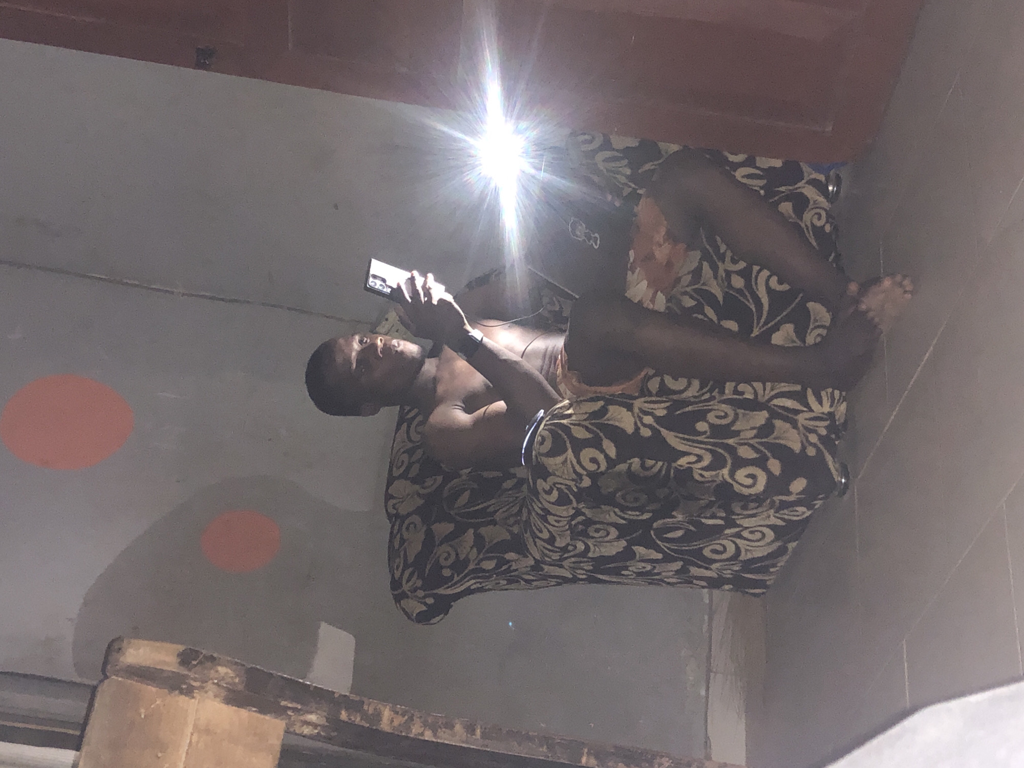A person is reclining on an ornate, patterned couch, angled diagonally across the image frame. The individual is looking at a mobile phone held in their hands. The person has dark skin and is shirtless. The bright reflection of a light source, presumably from a camera flash, washes out the details above the person. The background shows part of a wall with two circular, orange markings. A wooden beam is visible at the bottom of the frame. The location of the scene appears to be indoors. The city and state are unknown.