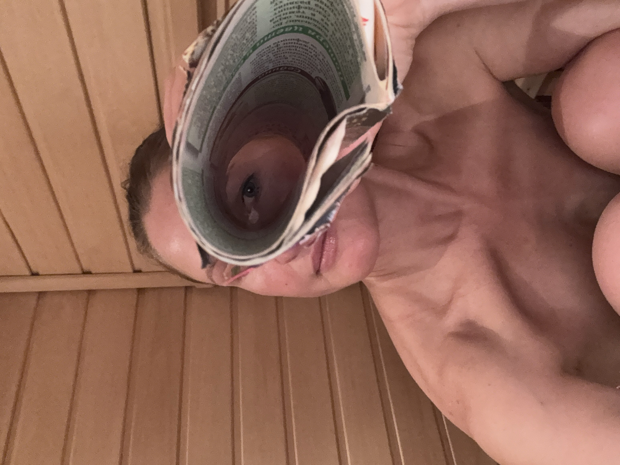 An intimate, overhead shot captures a person, likely a woman, lying down in an indoor setting. The backdrop consists of vertical wooden paneling, characteristic of a sauna or a similar wood-finished room, consistent with the location context of Chaikovsky, Russia. The person is engaged in a playful activity, looking through a tightly rolled-up newspaper or magazine, with one eye peering directly from the center of the roll. Her lips are also partially visible below the paper.
The rolled paper itself displays various printed texts, including green-colored sections and smaller black lettering. Faint Cyrillic characters are discernible on the paper, such as what appears to be "СООБЩЕНИЕ" (message/report) and "Гарантия" (Guarantee), further supporting the Russian context. The overall impression is one of relaxed amusement or curiosity, with the unique perspective created by the rolled paper suggesting a playful moment. The lighting is soft, highlighting the natural textures of the wood and the person's skin. No specific time of day or weather conditions are evident.