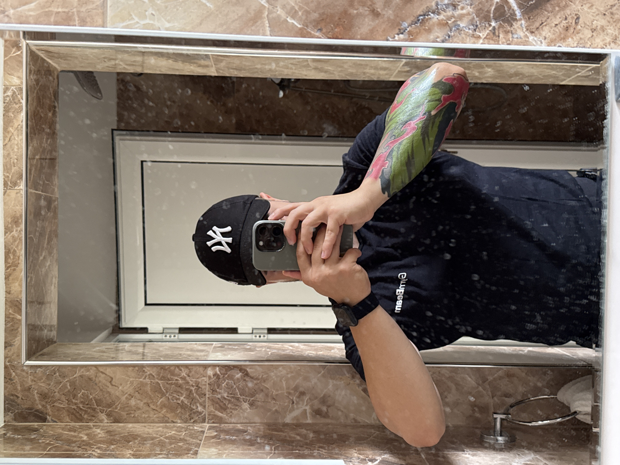 A person is captured in a mirror selfie, holding a smartphone with both hands to take the picture, obscuring their face. They are wearing a black baseball cap with a white "NY" (New York Yankees) logo and a dark, likely black, short-sleeved t-shirt. A black smartwatch is visible on their left wrist. Their right forearm is adorned with a vibrant, colorful tattoo featuring green, pink, and red patterns, possibly a full sleeve. The reflection reveals the setting to be indoors, likely a bathroom or hallway, characterized by polished brown marble-patterned tiles that frame the large mirror and cover the surrounding walls. The mirror itself shows signs of use, with numerous small water spots or dust particles on its surface. Reflected in the background, a white-framed window is visible, along with a chrome towel rack holding a white towel in the lower right corner. Text on the person's shirt is visible in reverse, appearing as "m99BmЭ" which suggests "GymBeam" or similar branding.