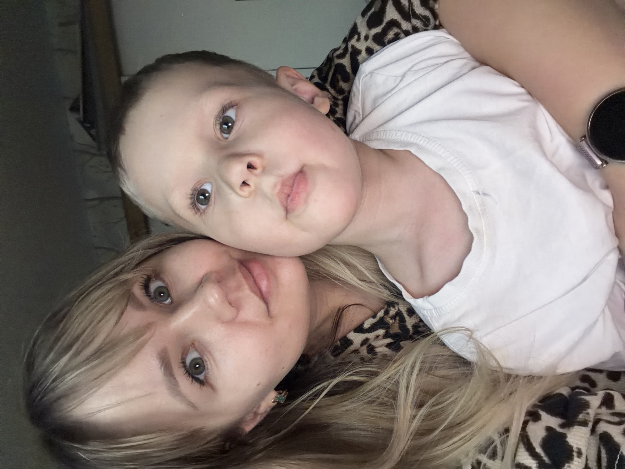 This media file presents a close-up, rotated portrait of an adult female and a young child, likely taken as a selfie indoors. The image is oriented 90 degrees clockwise, depicting the subjects lying on their sides.
In the lower half of the frame, the adult female is visible with long, wavy blonde hair, light skin, and light-colored eyes (appearing greenish-grey). She has a calm expression with slightly parted lips and looks directly at the camera. She appears to be wearing a white top, and a leopard print fabric is prominent around her, possibly part of her clothing or a blanket. A small, possibly jeweled earring is visible in her left ear, and a silver smartwatch is on her right wrist.
Above her, resting his head against hers, is a young child, likely a boy. He has light brown or blonde hair, light skin, and light-colored eyes (appearing brownish-grey). He is wearing a plain white short-sleeved top. His expression is neutral, with his lips slightly pursed, and he is also looking towards the camera.
The subjects are in close, affectionate contact, suggesting a tender moment. The background is dark and out of focus, indicating an indoor setting, possibly a bed or couch, with subdued lighting. The exact time of day or specific location is not discernible beyond it being an indoor residential environment. There is no visible text in the image.