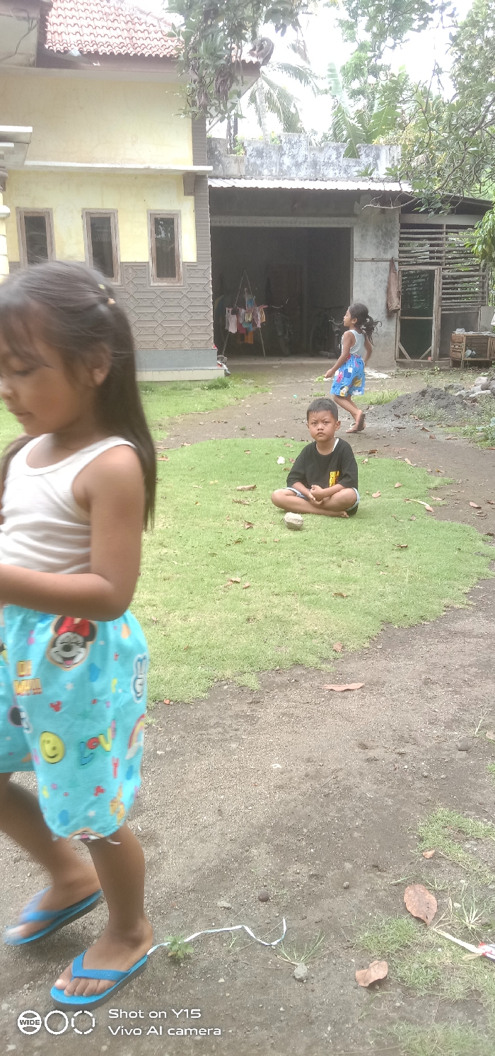 The image depicts an outdoor scene in a residential area of Ranugedang, Indonesia, featuring three children. In the immediate foreground, a young girl with long dark hair, wearing a white sleeveless top and light blue shorts adorned with cartoon characters and emojis, is partially visible on the left, moving towards the right. She wears blue flip-flops.
In the midground, a boy with dark hair and a neutral expression sits cross-legged on a patch of green grass, facing the viewer. He is dressed in a black t-shirt with light-colored text and dark shorts. A small, light-colored stone or object rests on the grass directly in front of him. Behind him, towards the right, another young girl is seen walking away from the camera and towards the right side of the frame. She wears a light-colored sleeveless top and patterned shorts, and her dark hair appears to be in motion.
The ground consists of irregular patches of vibrant green grass interspersed with areas of bare, light brown dirt and small gravel. Scattered dried leaves are visible on both the grass and dirt. In the background, on the left, stands a two-story house with a light yellow facade, brown-framed windows, and a lower section featuring a patterned grey and brown textured wall. Its roof is tiled. To the right of this house, a single-story concrete structure, likely a garage or utility building, has an open entrance revealing a dark interior where colorful clothes are hanging on a rack, and a bicycle is partially visible. This structure is covered by a corrugated metal roof. Lush green foliage and various trees, including what appears to be a palm tree, fill the upper background, indicating a tropical environment. The scene is illuminated by natural daylight, suggesting an overcast or diffused light condition.