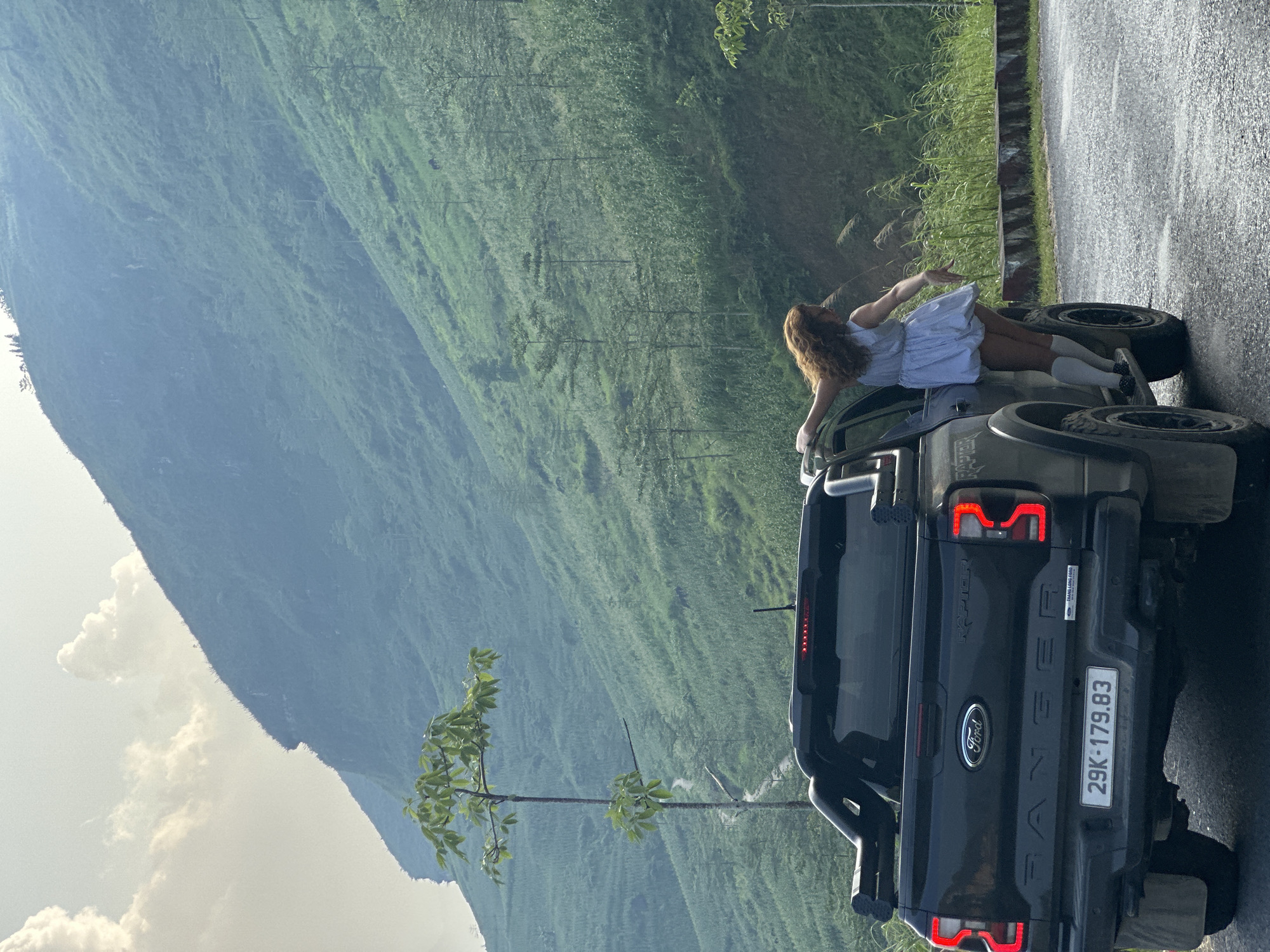 The image shows a woman leaning on a black Ford Ranger pickup truck on a road in a mountainous area. The license plate of the truck reads "29K-179.83". The background features a large, green, mountainous landscape. The woman is wearing a light-colored dress and white socks. The road is wet. A small tree grows beside the truck. The location is Du Già Commune, Vietnam.