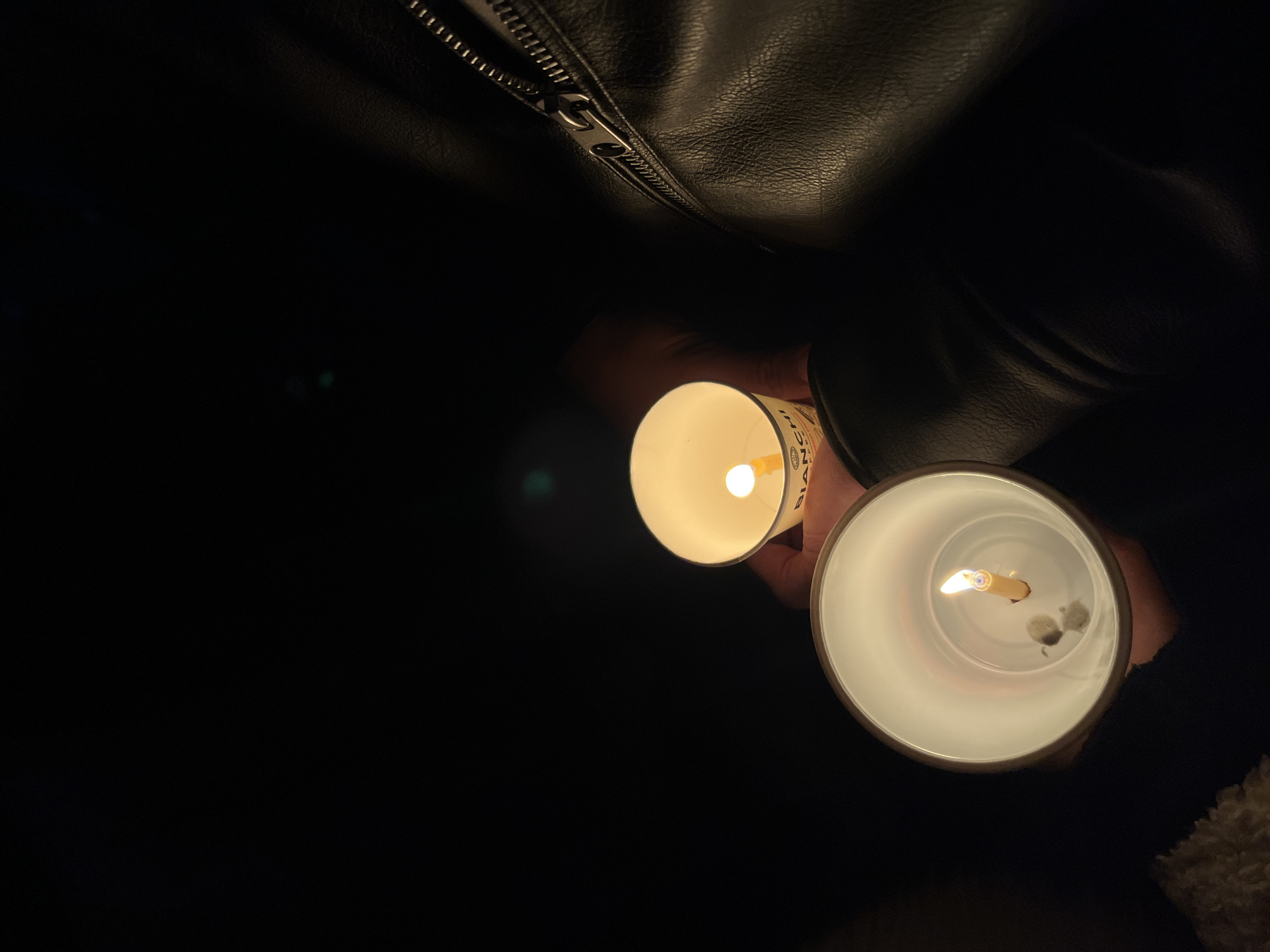 A person is holding two lighted white votive candles in the dark. The candles are in white paper cups, and the person is wearing a black leather jacket with a visible zipper. The background is mostly dark, with a few hints of the surroundings. There is no information available to determine that this image was taken in Gotse Delchev, Bulgaria.