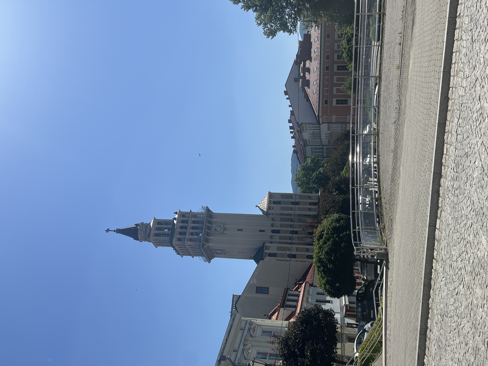 The media file displays an outdoor scene with a prominent bell tower dominating the left side of the frame. The bell tower is a tall, slender structure with multiple levels featuring arched openings and a dark, pointed spire topped with a cross. A clock is visible on one of the tower's faces. Adjacent to the tower are buildings with red tile roofs and multiple windows.
In the foreground on the right, a paved walkway made of grey interlocking stones leads the eye into the scene. A modern metal railing with clear panels separates the walkway from a lower area where cars are parked. Lush green trees are scattered throughout the image, with one particularly large tree canopy visible at the top left. The sky is a clear, bright blue, suggesting a sunny day. A few birds are visible as small dark specks against the sky, one near the bell tower and another further left. The scene appears to be a town square or a street in a European city, bathed in bright sunlight, indicating it is daytime, likely late morning or early afternoon. The overall atmosphere is tranquil and bright. No visible text is present in the image.