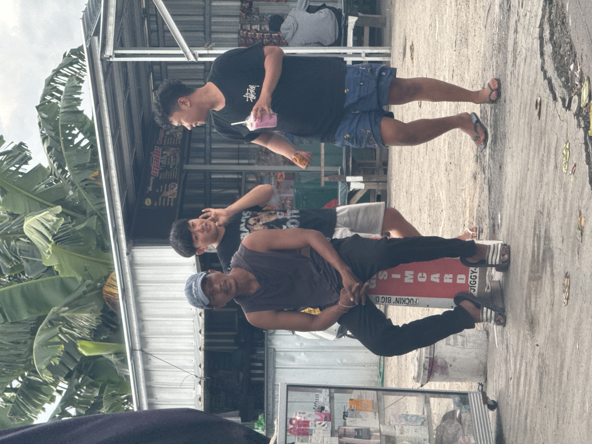 The image is of a street scene in North Kuta, Indonesia. There are three males present. One is older, wearing a grey tank top and navy blue cap, black pants and sandals, and leaning against a red sign with writing on it. A second male stands behind and to the right, wearing a black t-shirt with a white graphic, grey shorts, and sandals. His left hand makes a gesture near his face. The third male, standing toward the left and at the top of the frame, wears a black t-shirt and blue patterned shorts, holding a pink drink in his left hand and some food in his right. A glass display case stands in front of the older male, and a metal-roofed structure looms in the background. To the left are large banana trees. The ground is concrete and appears somewhat dirty.