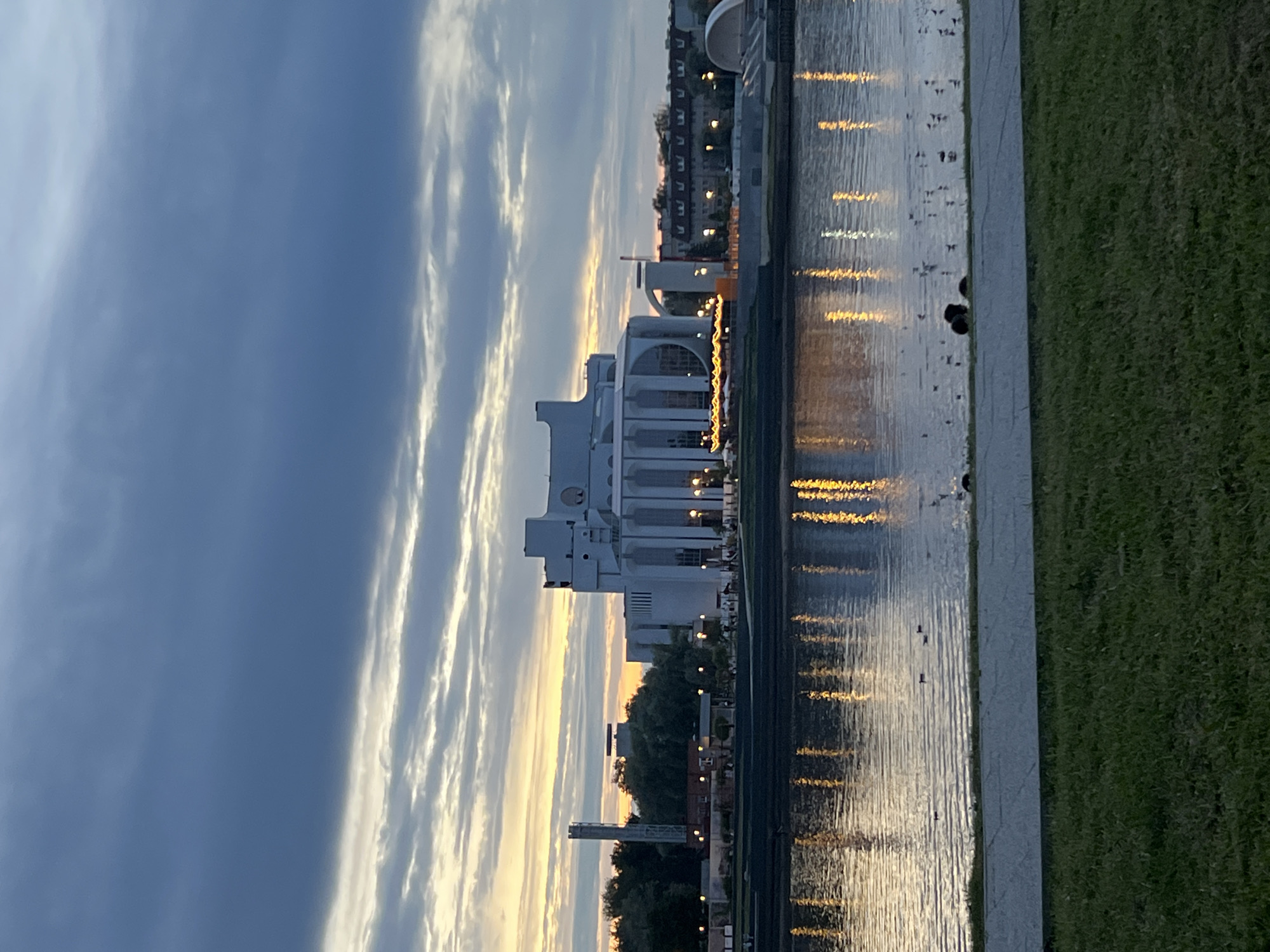 The image shows a wide river in Veliky Novgorod, Russia, reflecting the lights of buildings and the sky at dusk. On the far bank of the river, a large, modern white building with tall columns and arched windows is visible, likely a cultural center or theater. The sky above is cloudy with streaks of orange and yellow from the setting sun, and the clouds are dark blue and grey. The river's surface is rippled, with the reflections of lights creating a shimmering effect. In the foreground, there is a grassy area and a paved walkway. Several ducks and other birds are present on the water and near the shore. The overall scene conveys a sense of tranquility and urban beauty at twilight.