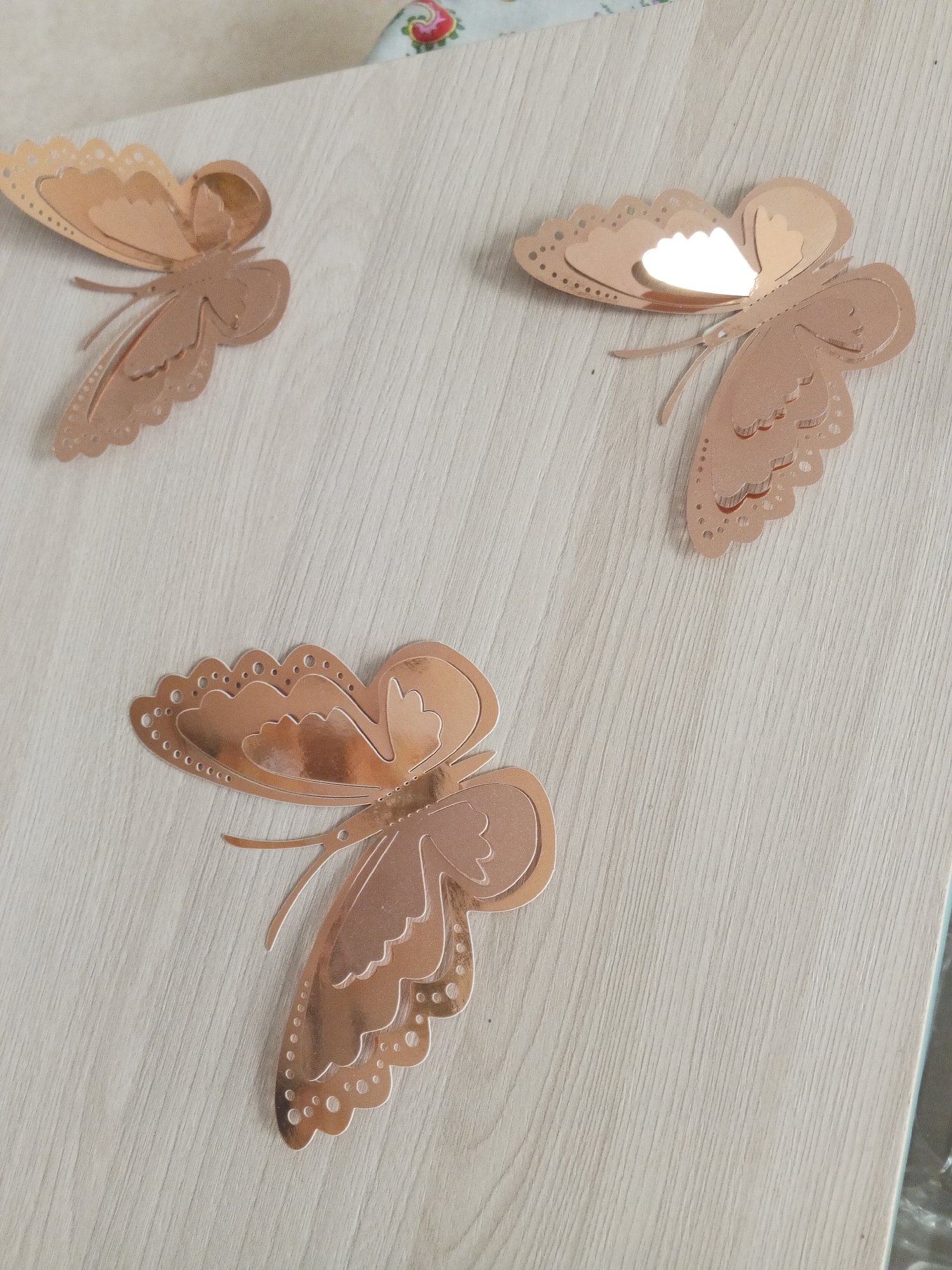 Three rose gold butterfly decorations are affixed to a light-colored wooden surface. The butterflies are decorative and appear to be made of a metallic material. They are positioned in a scattered arrangement across the surface. The top left butterfly is angled downwards and to the right, showing its patterned wings. The top right butterfly is facing towards the viewer and slightly upwards, with a strong reflection on its wings. The bottom butterfly is the largest and is positioned horizontally, extending from the left to the center of the frame, also displaying its detailed wings and body. The wooden surface has a visible grain pattern. No people or any other objects are present in the scene. This image was taken in Danilov, Russia.