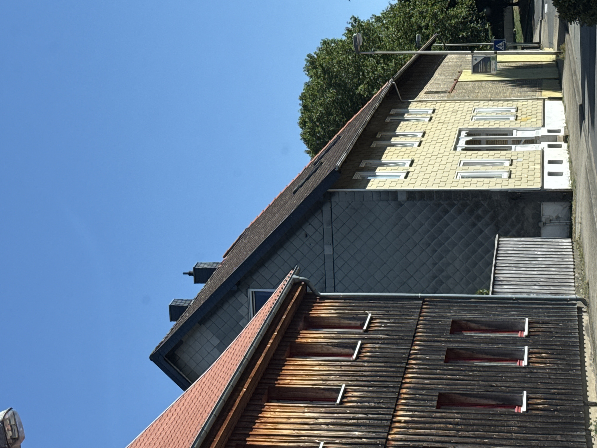 The image shows a street scene in Herzberg, Germany, featuring two adjacent buildings under a clear blue sky. The building on the left is a wooden structure with a red-tiled roof and vertical planks, showing three tall, narrow windows with red frames and white sills. The building to its right is a multi-story structure with a dark gray, diamond-patterned facade on its upper section and a light yellow, shingled facade on the lower two stories. This building has multiple white-framed windows and a prominent white door with a glassed-in porch. A paved sidewalk runs in front of the buildings, and a street is visible to the right, with a lamppost and a pedestrian crossing sign. A dense green tree line forms the background.