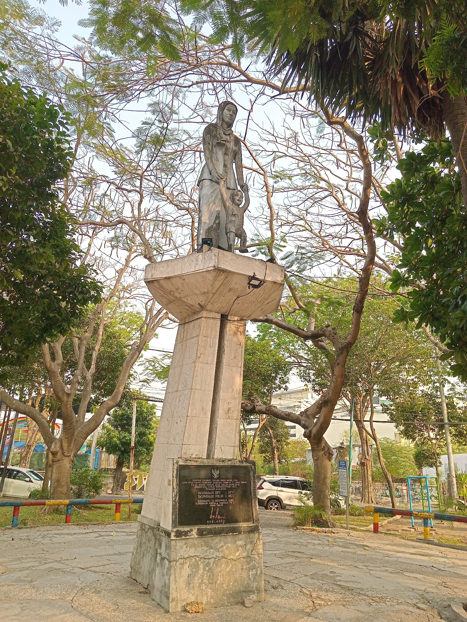 This is a full-shot, eye-level, outdoor photograph of a statue in Randuagung, Indonesia. The statue depicts a woman holding a child's hand. The monument is made of stone and sits on a concrete pedestal. A plaque with text in Indonesian is affixed to the front of the pedestal. Surrounding the statue are several trees with bare branches and green foliage, casting shadows on the ground. In the background, there are buildings, a white car, a playground with colorful bars, and a blue and yellow fence. The sky is visible in the upper part of the frame. The lighting suggests it is daytime, likely late afternoon due to the warm tones.